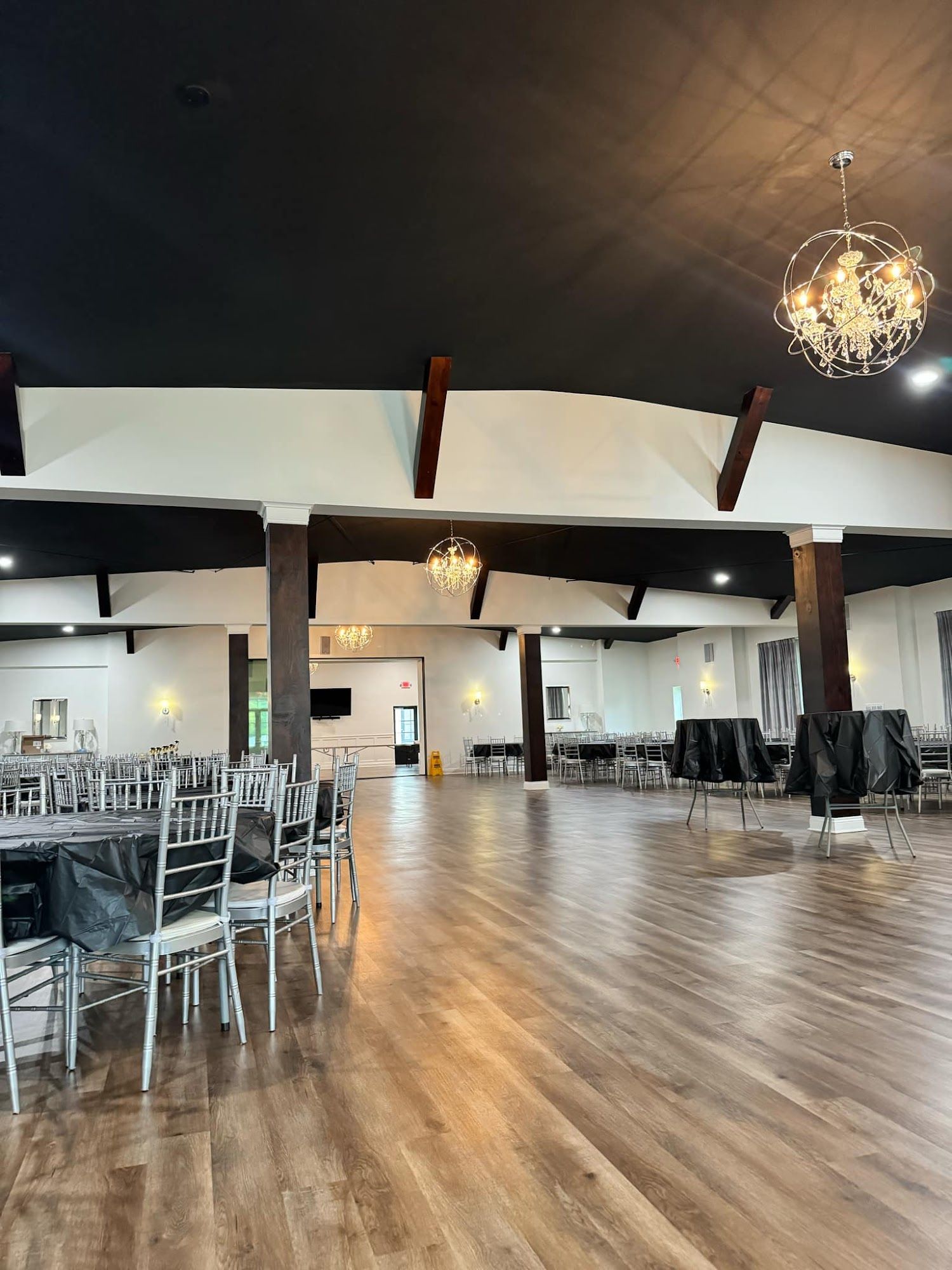 Large event hall with wooden floor, tables, chairs, and chandeliers. Black ceiling and white walls.