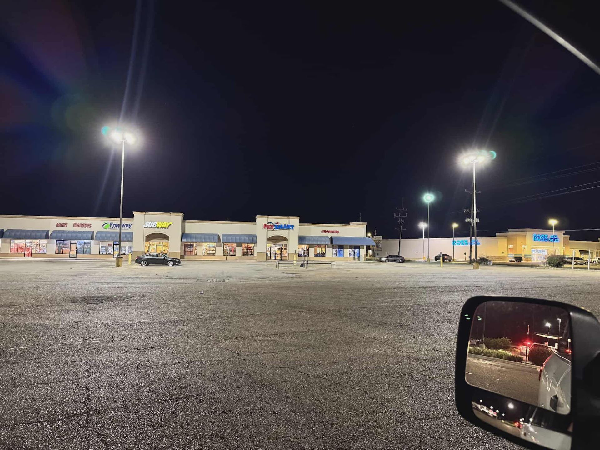 Night view of a mostly empty parking lot in front of a strip mall with bright overhead lights.