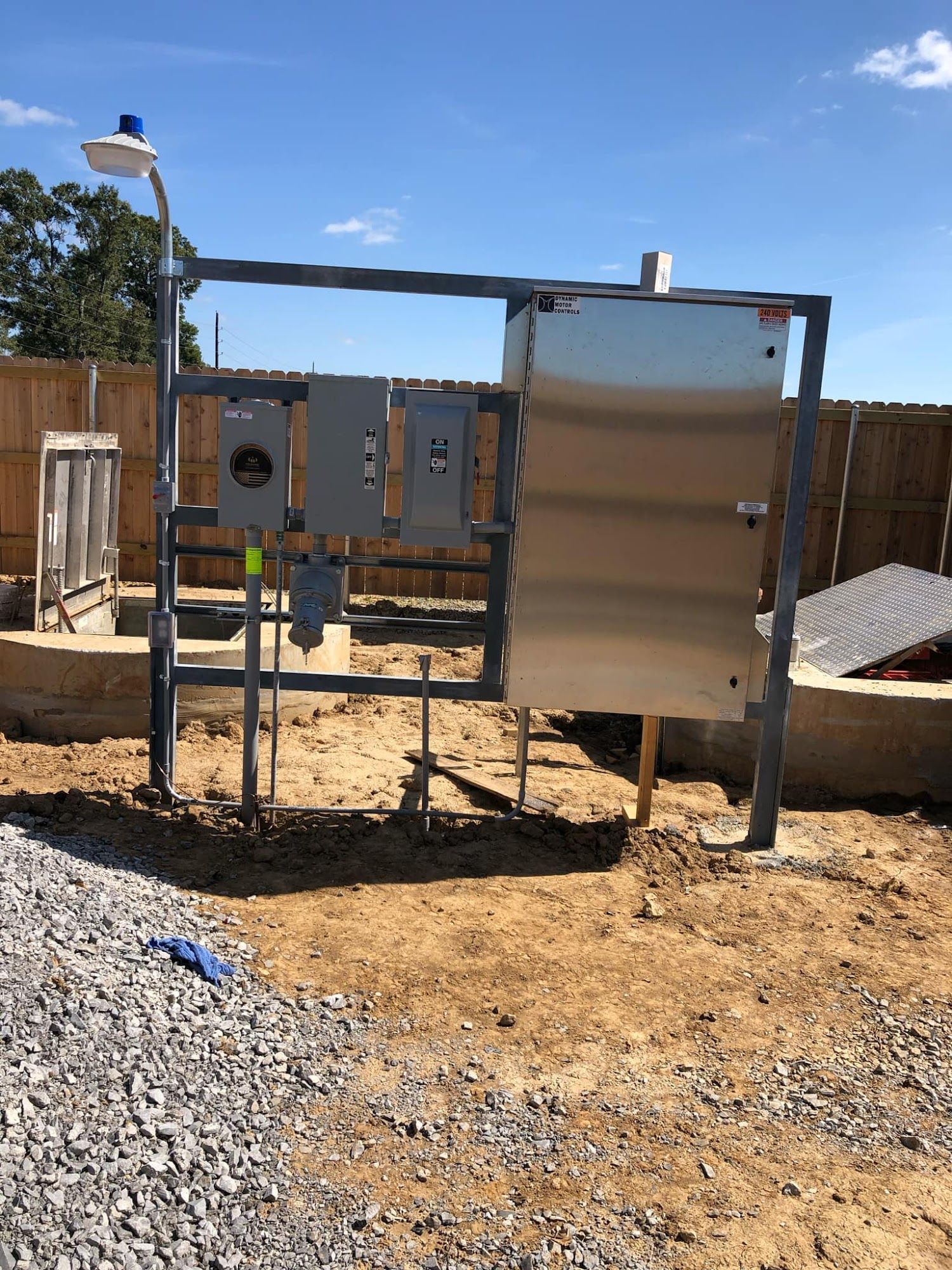 Outdoor electrical panel and framework on dirt, with blue light and surrounding fence.