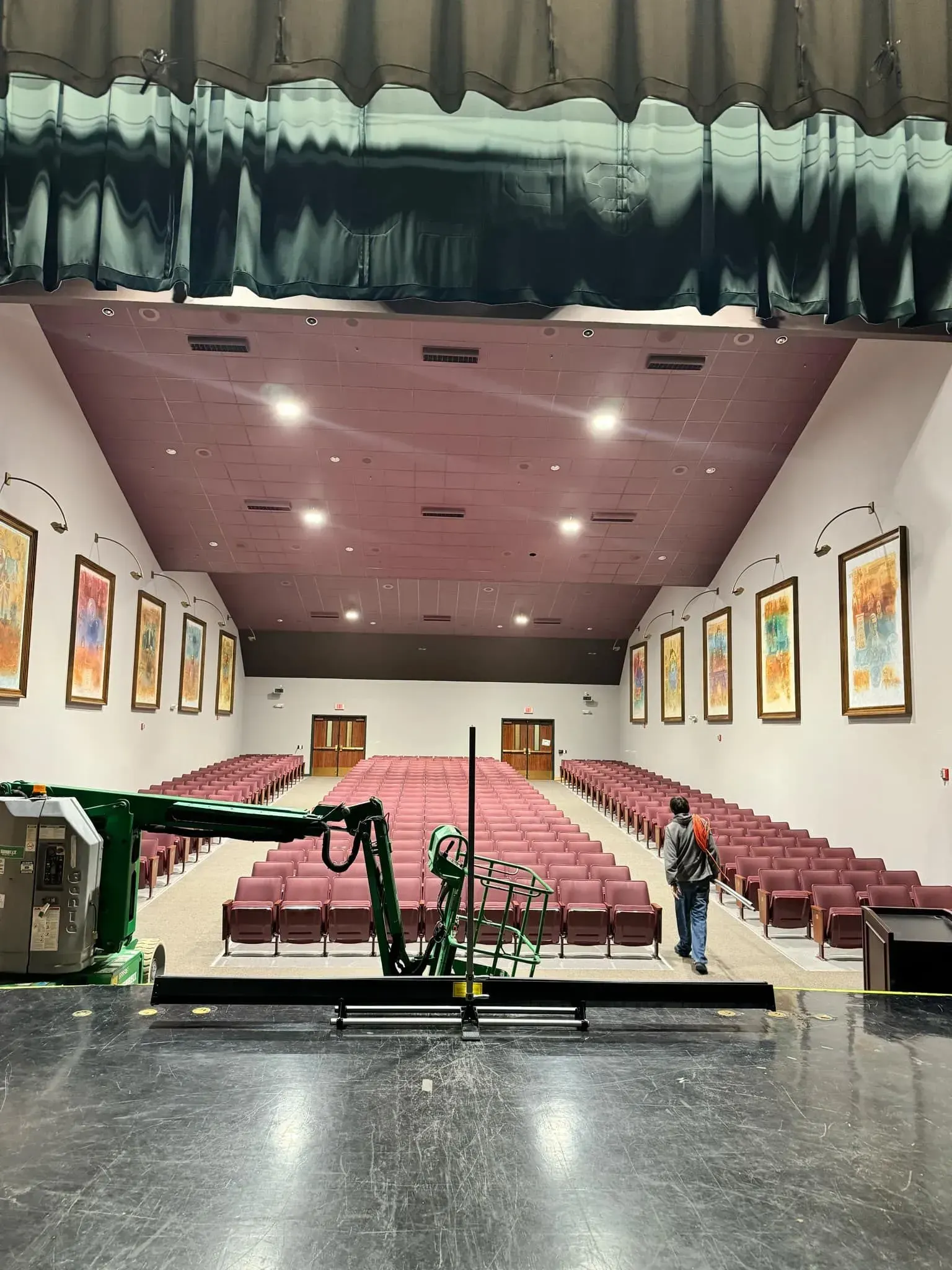 Interior view of a maroon-seated auditorium from the stage, with a person standing by a floral display.