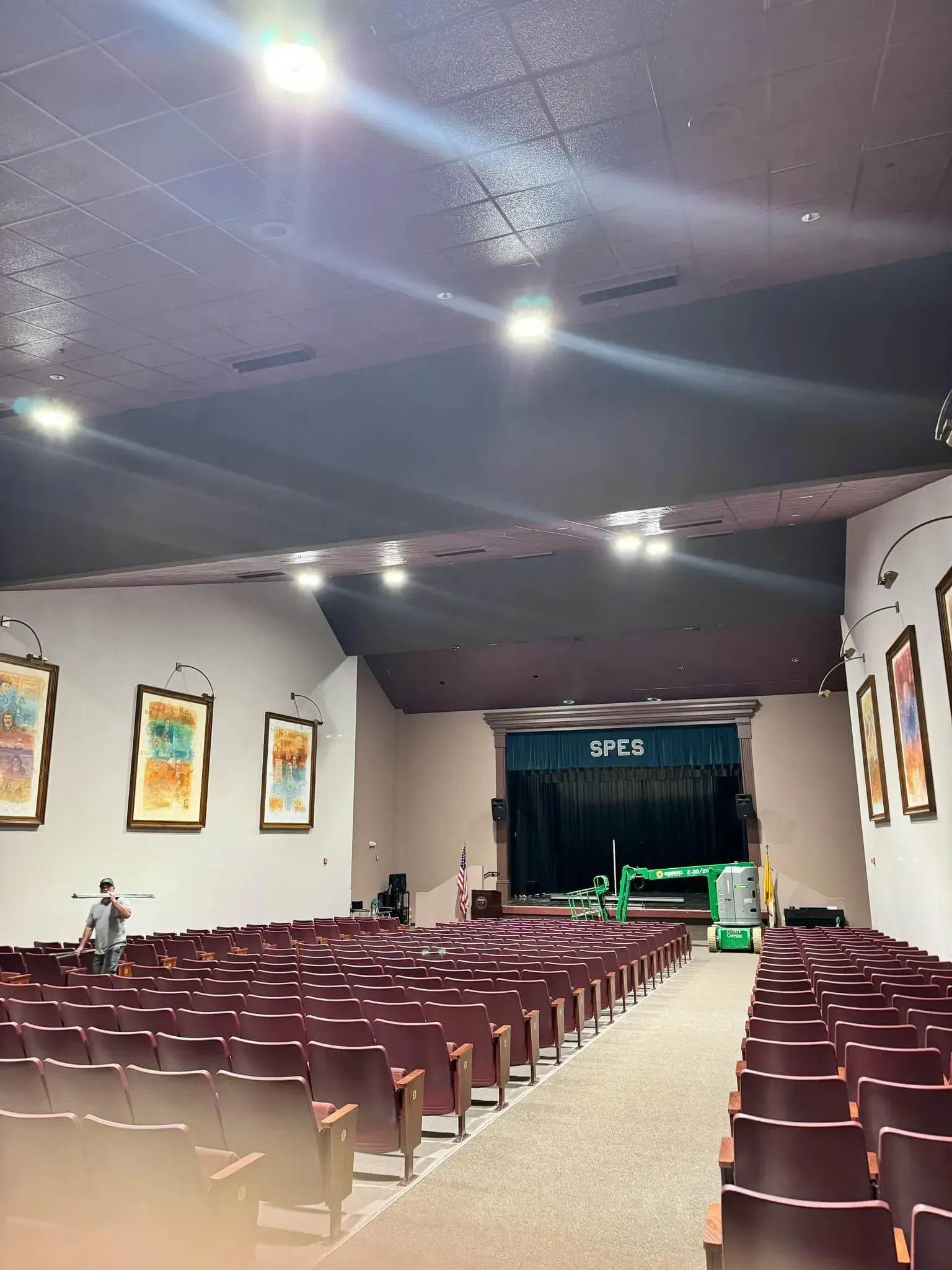 Auditorium interior with rows of maroon seats, stage, paintings on walls, and work being done.