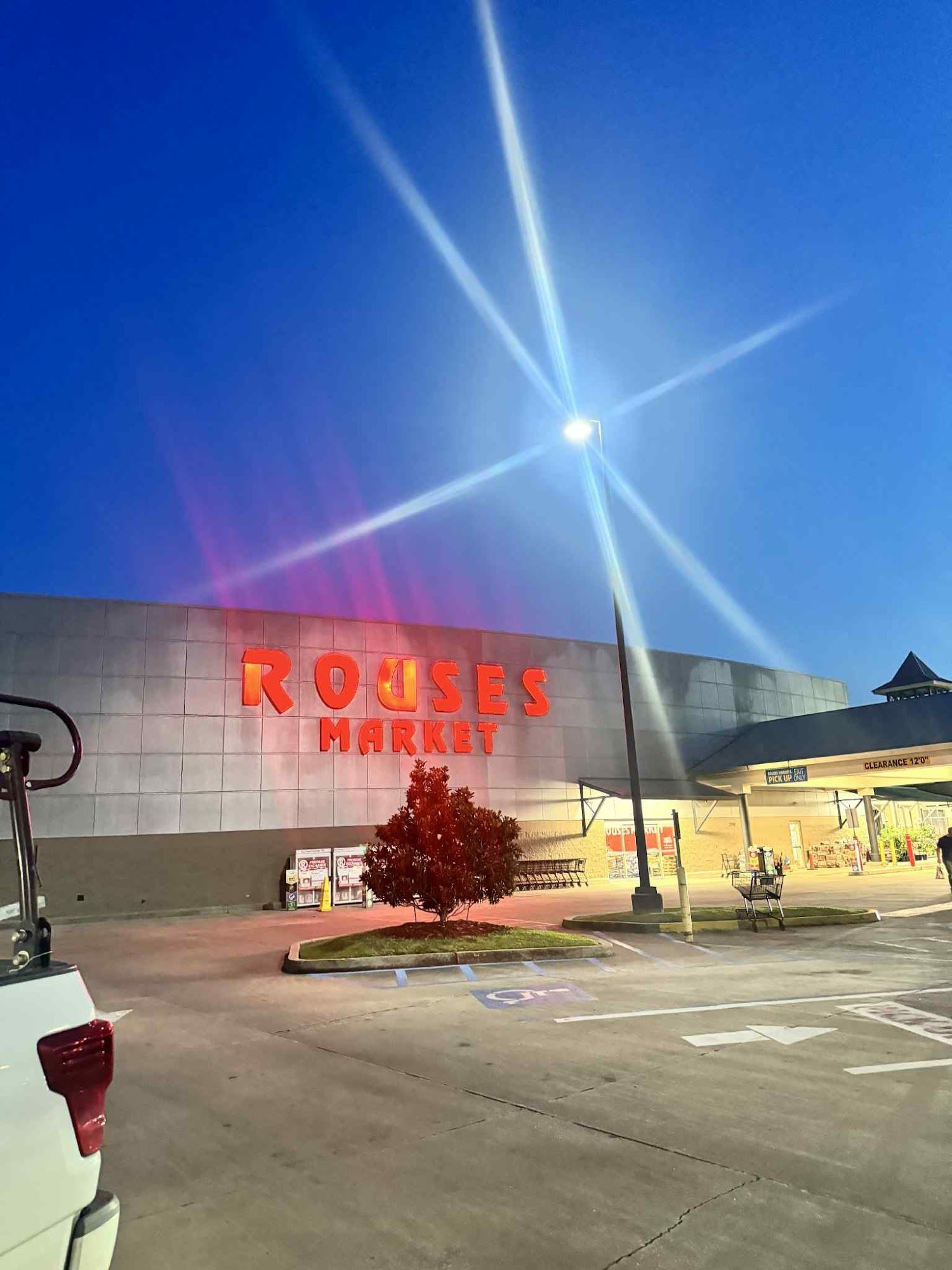 Rouses Market exterior at dusk; red sign on gray building; bright street light flares in sky.