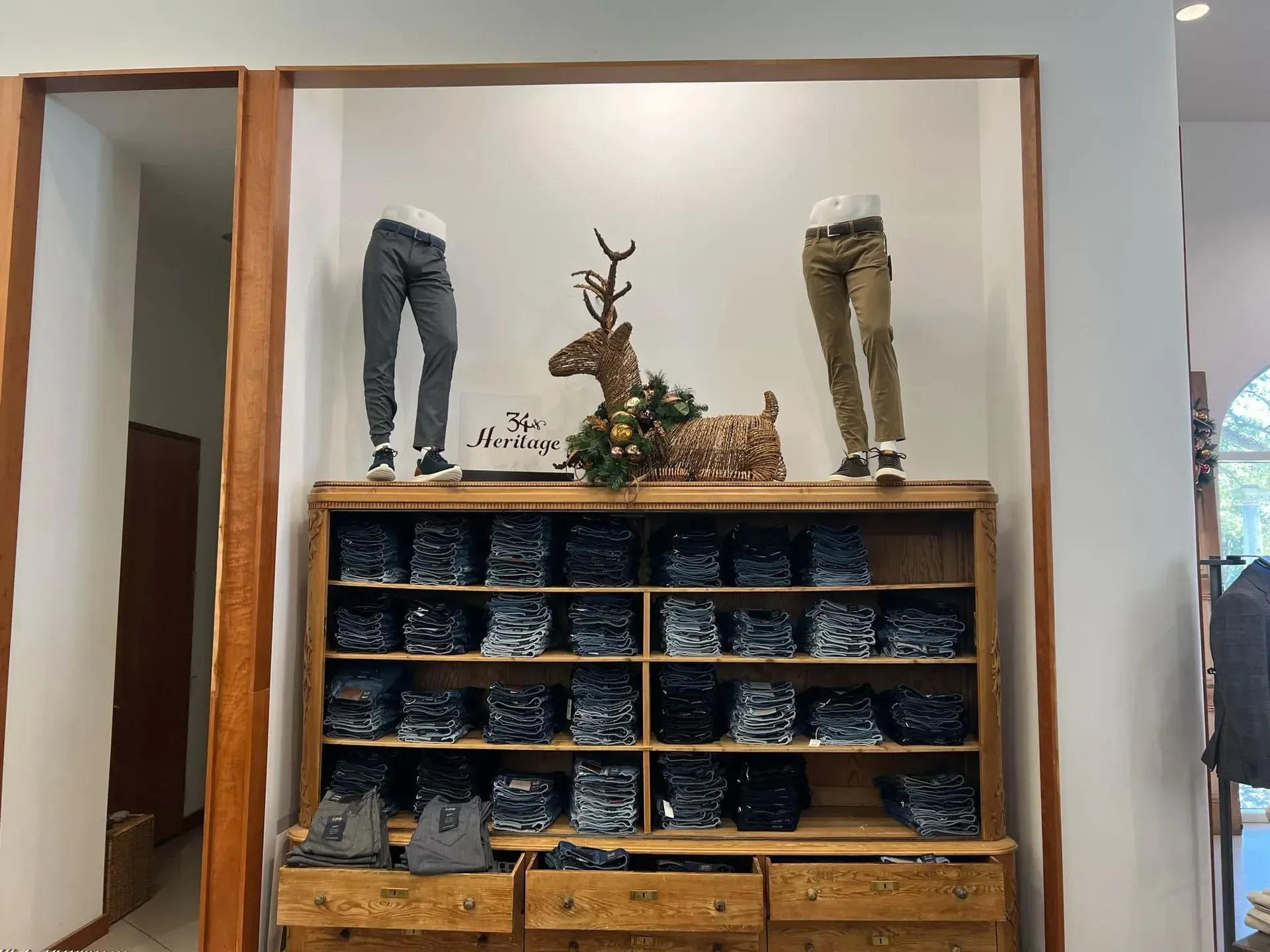 Clothing store display: jeans in cubbies, two mannequins wearing pants, a deer statue on a wooden cabinet.