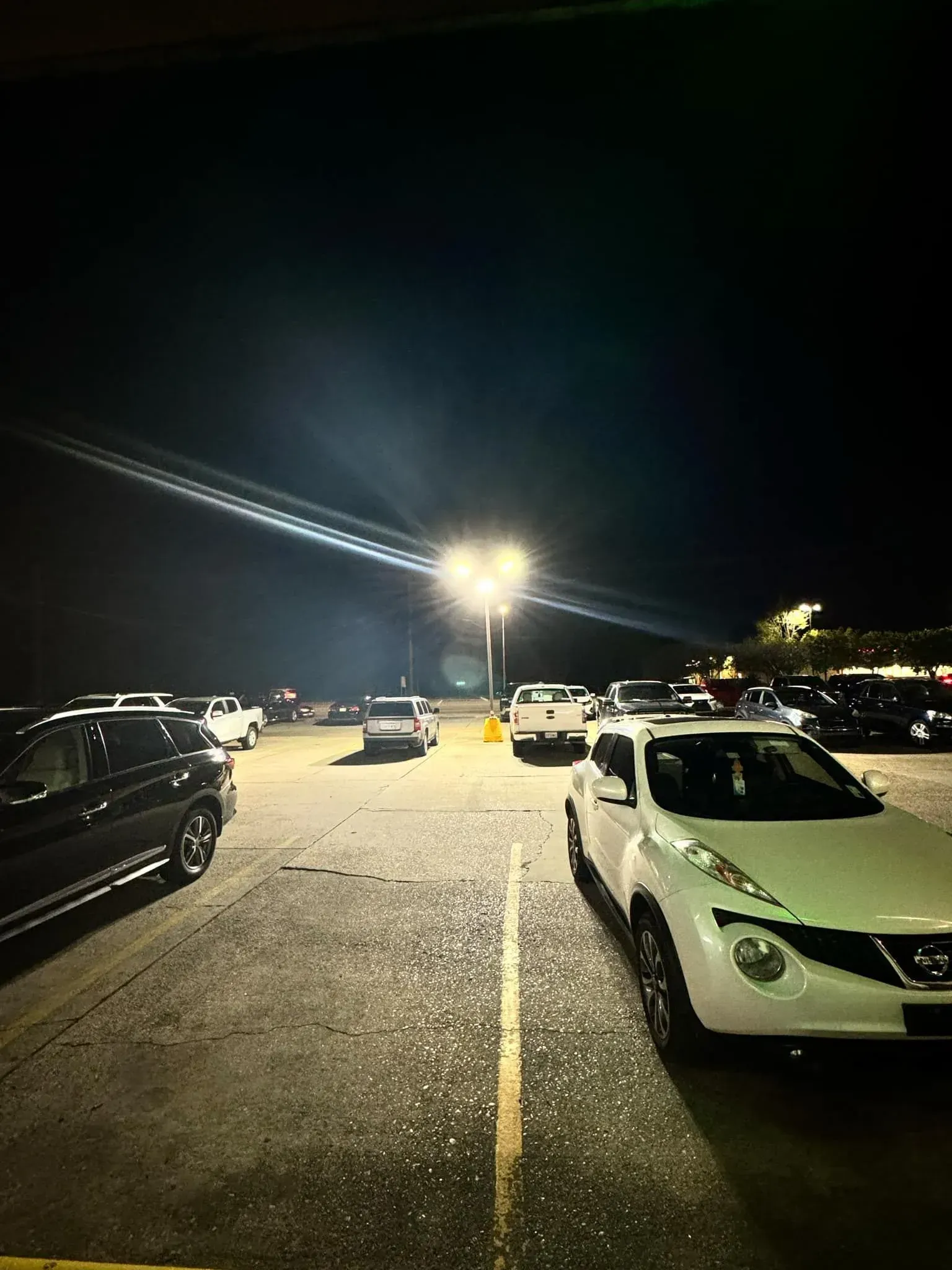 Nighttime parking lot with cars, bright overhead lights, and a white SUV in the foreground.