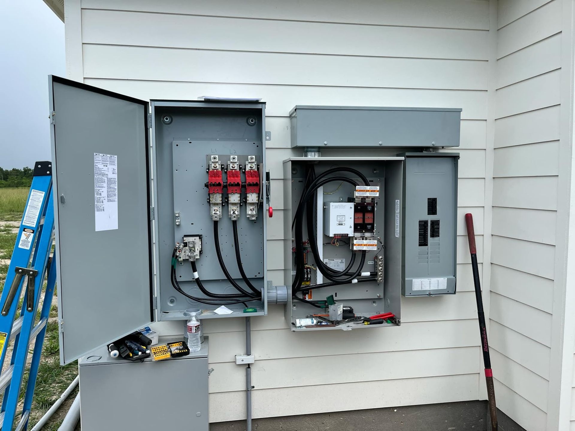 Electrical junction boxes mounted on a white house exterior with open panels revealing wires and components.