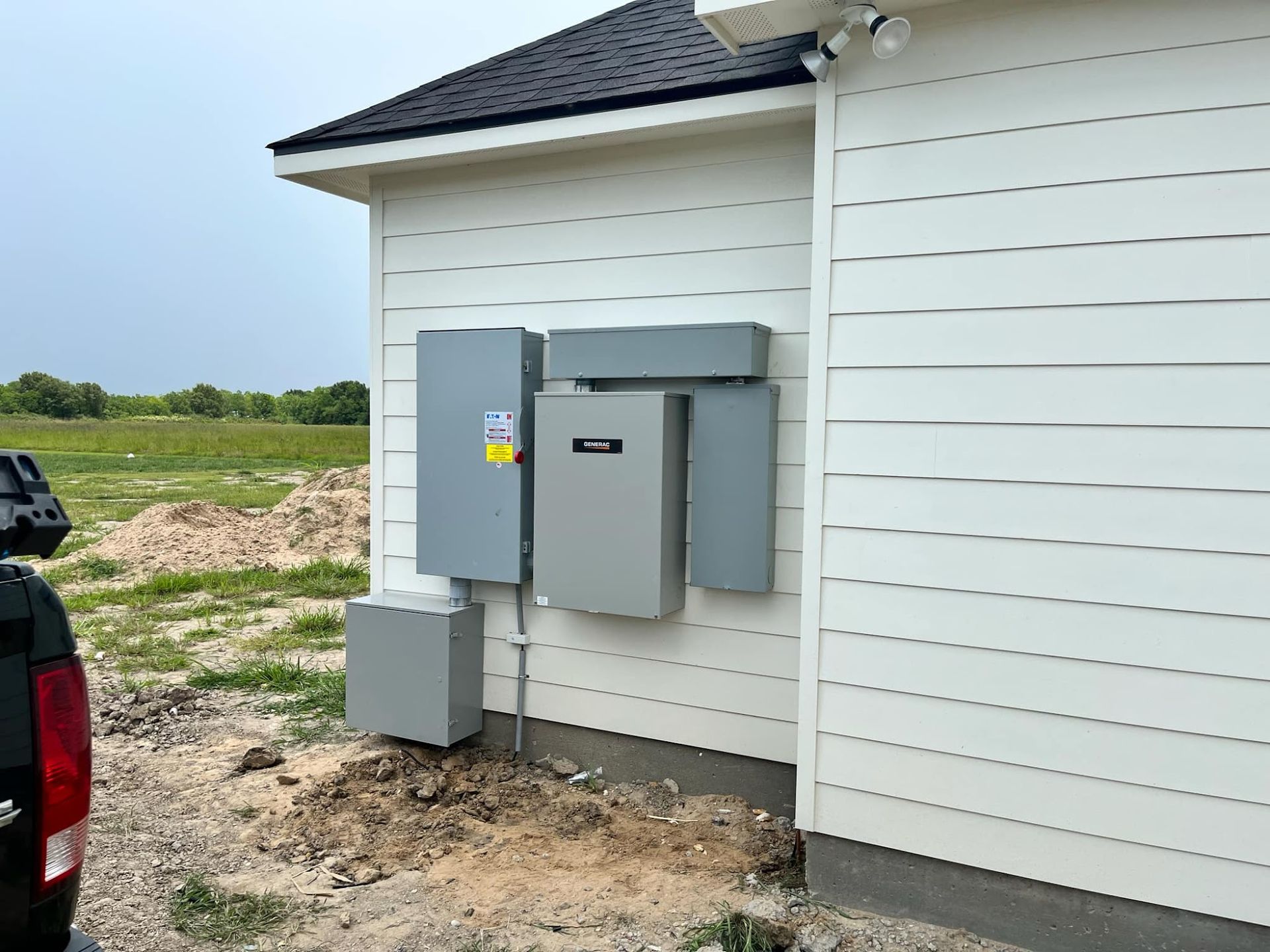 Gray electrical boxes mounted on the white siding of a house with a black roof.