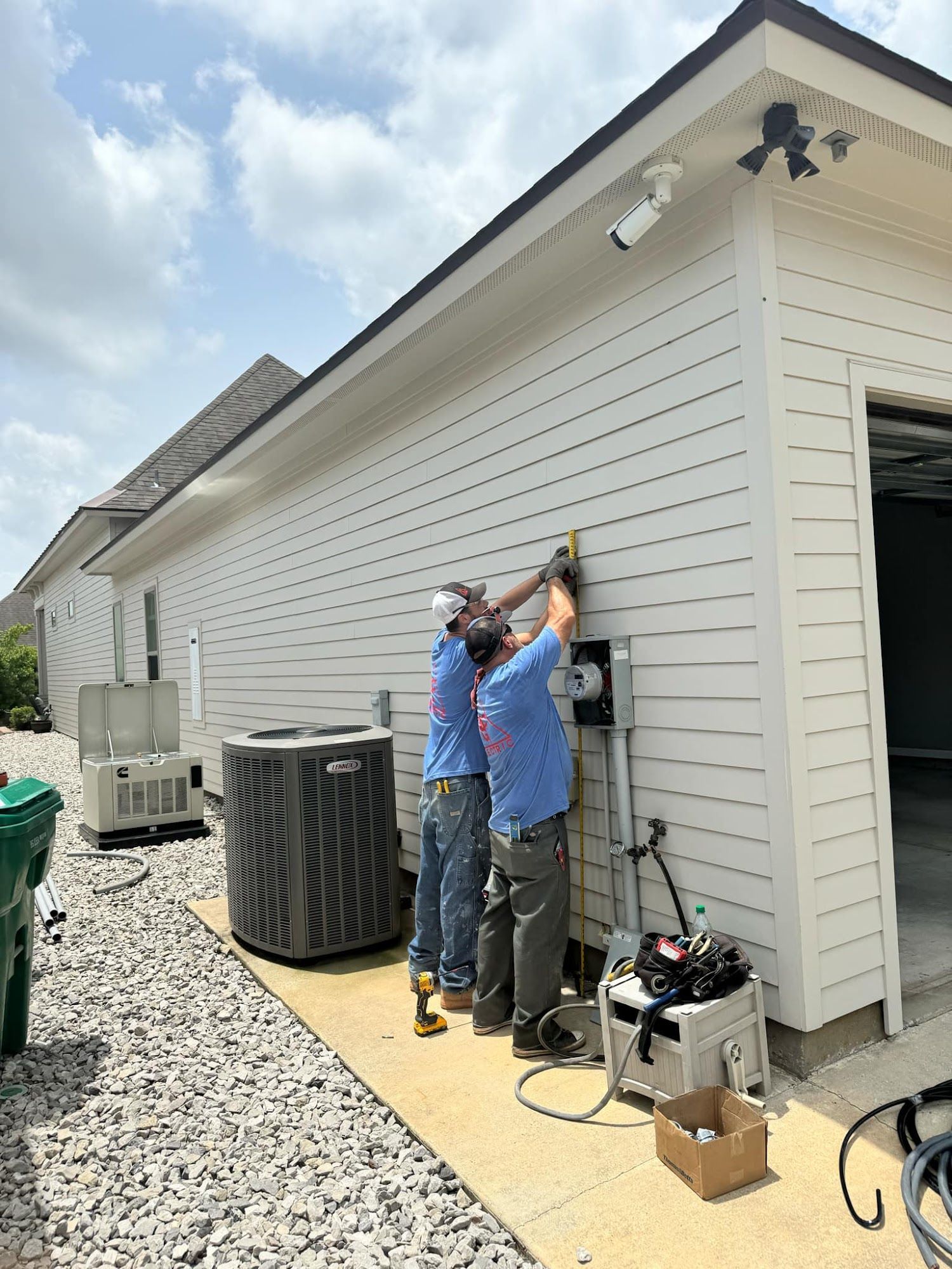 Two electricians work on a gray electrical panel on a white house exterior near an AC unit and garage.