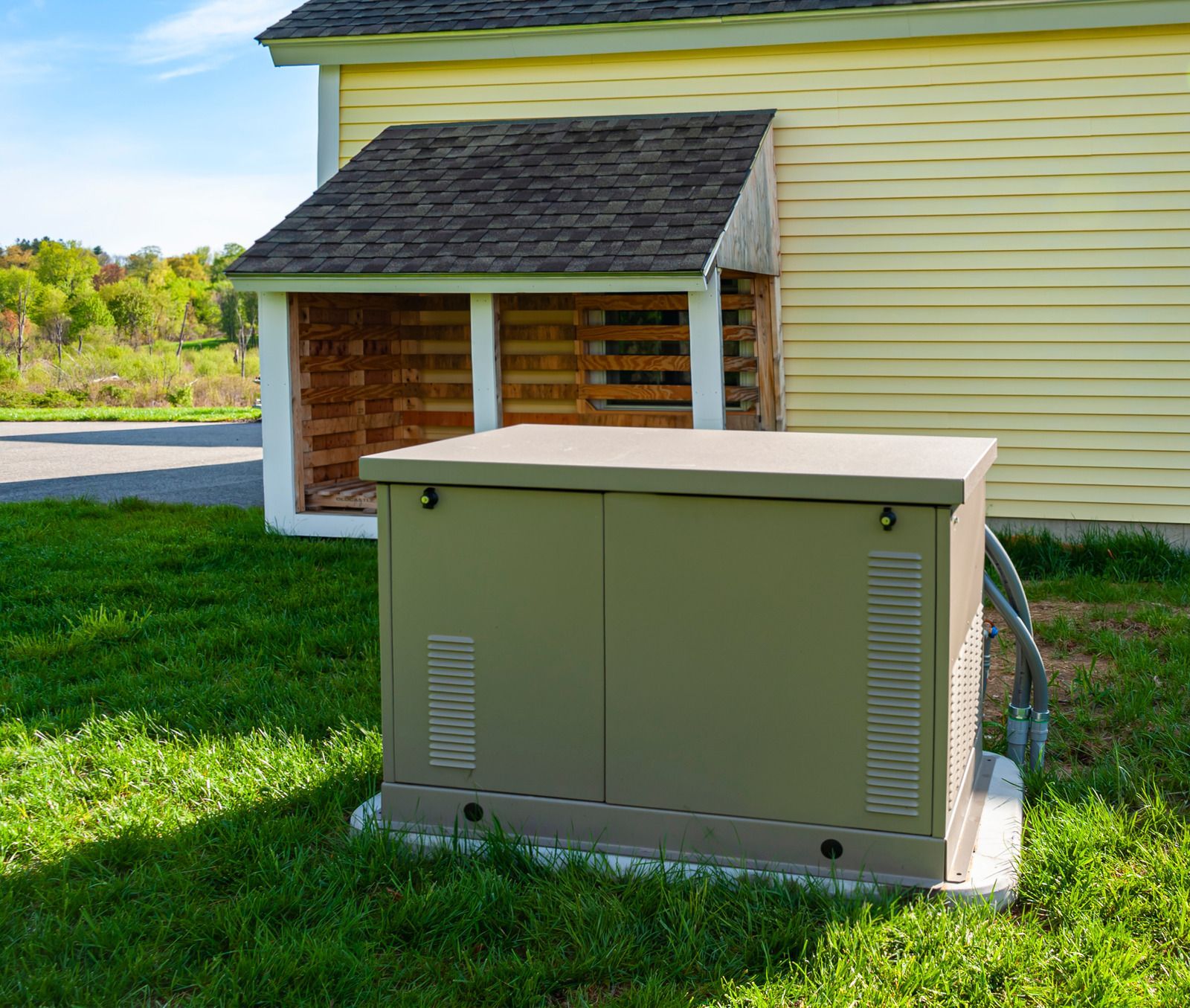 A generator on a concrete pad near a yellow house with a wood storage area in a grassy yard.