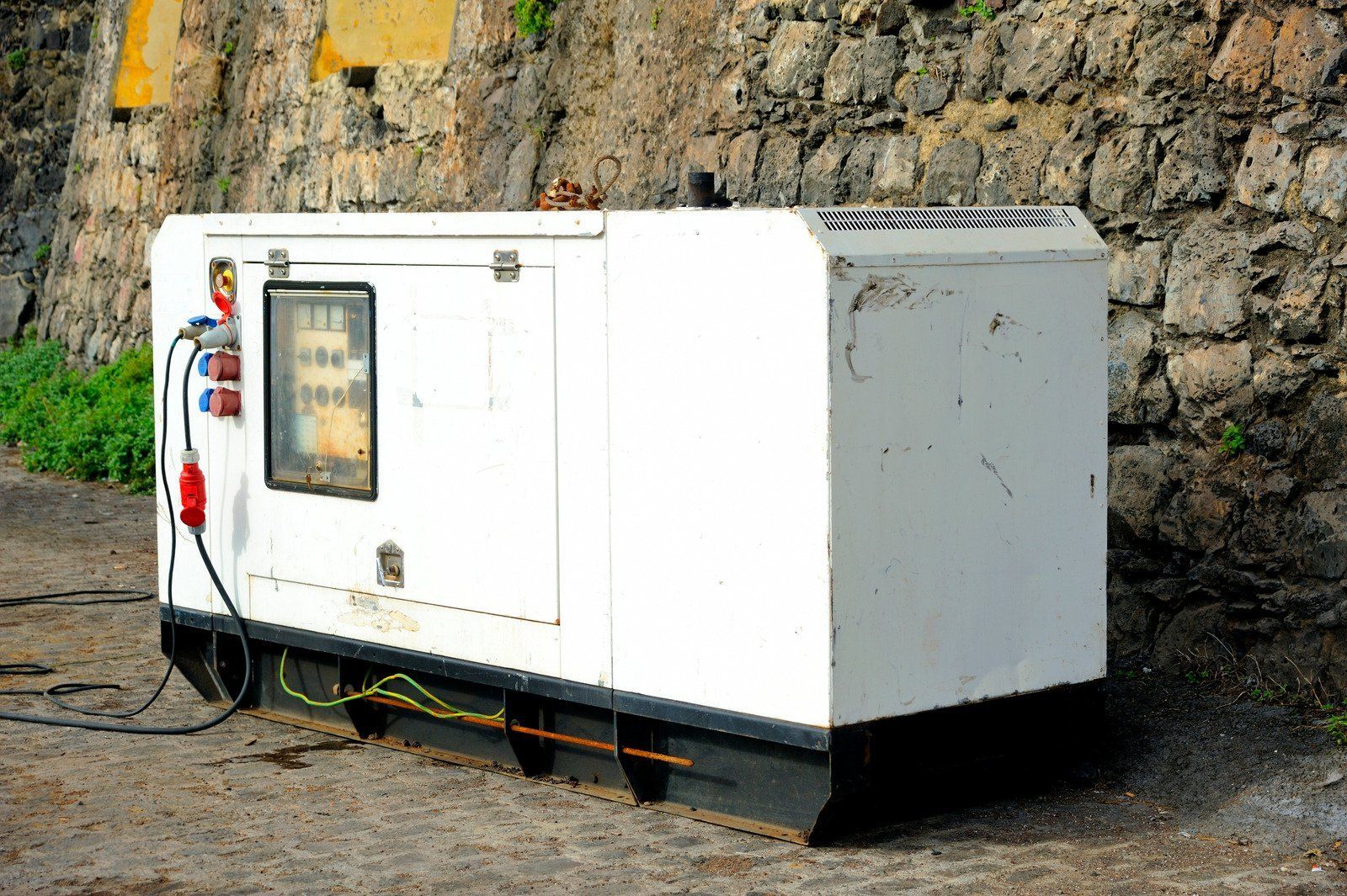 White generator with black base, against a stone wall. Electrical cords connected.