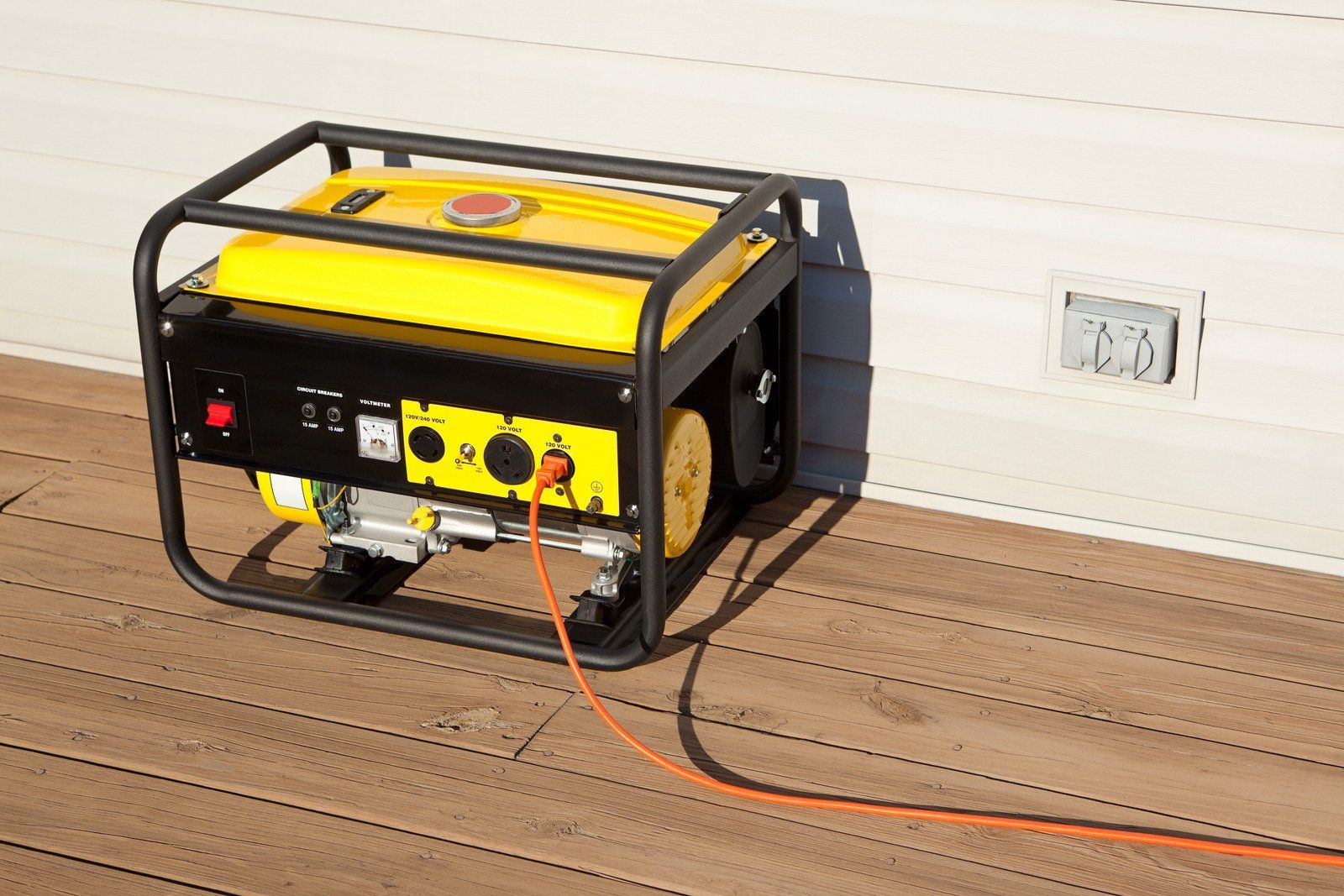 Yellow generator on a wooden deck, connected to a wall outlet via an orange cord.