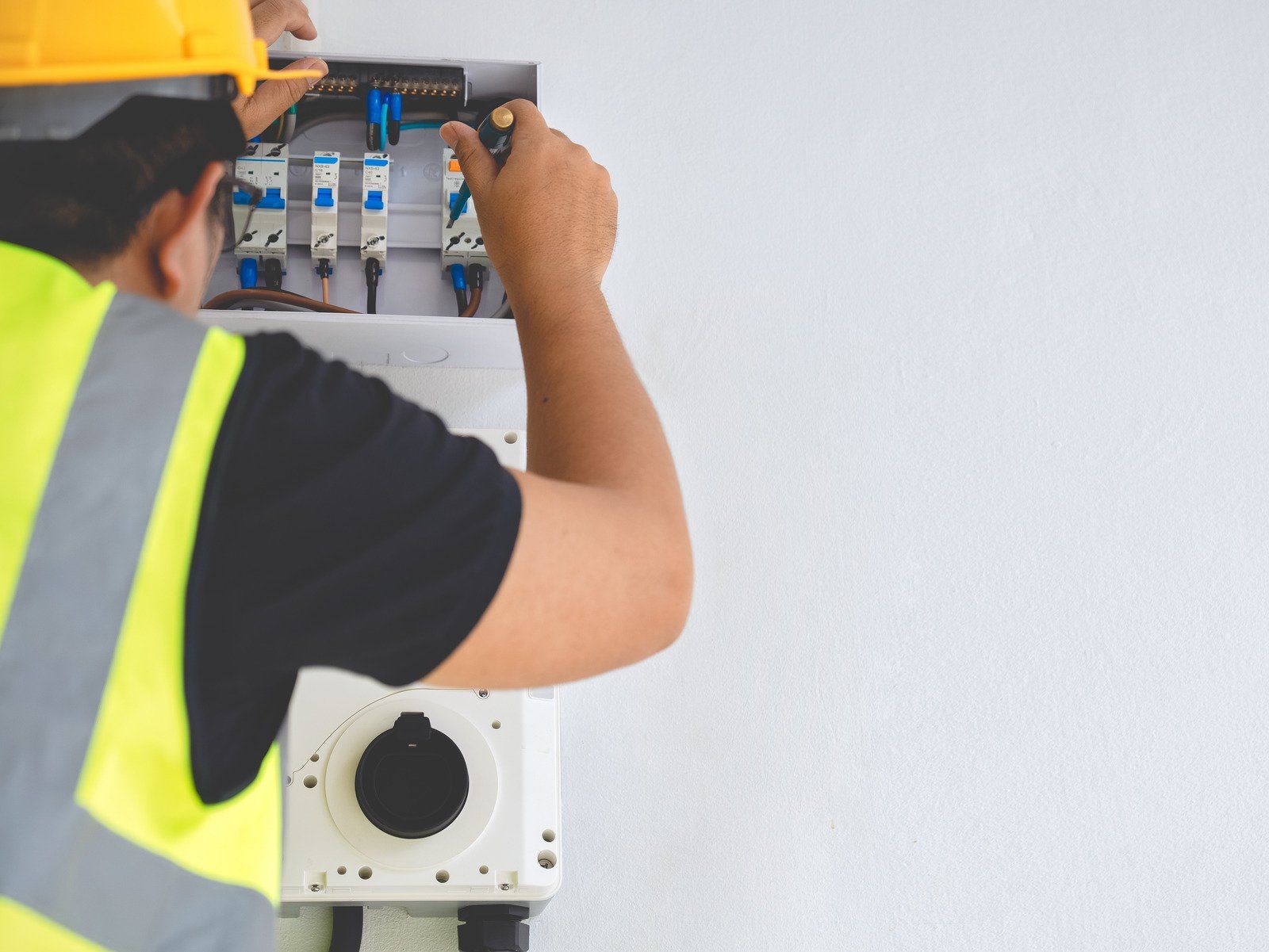 Electrician in yellow vest and hard hat working on an electrical panel against a white wall.