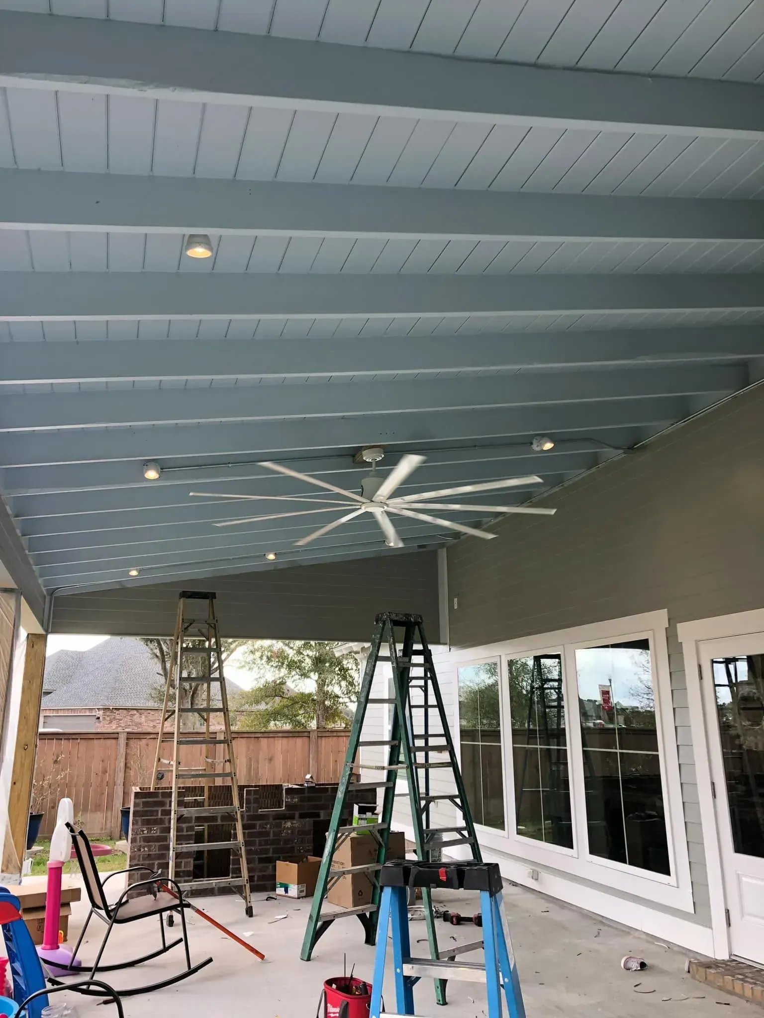 Patio ceiling with painted blue planks, lights, and a large fan; ladders and tools are present.