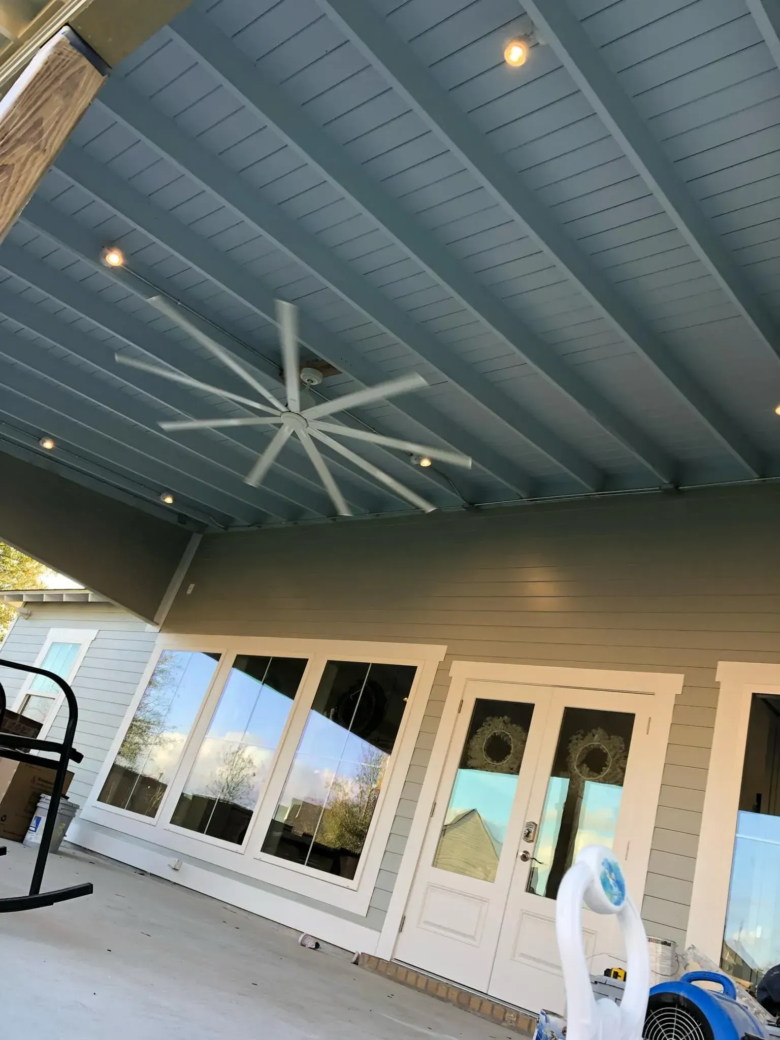 A covered patio with a large ceiling fan and recessed lighting; blue ceiling, and gray exterior.