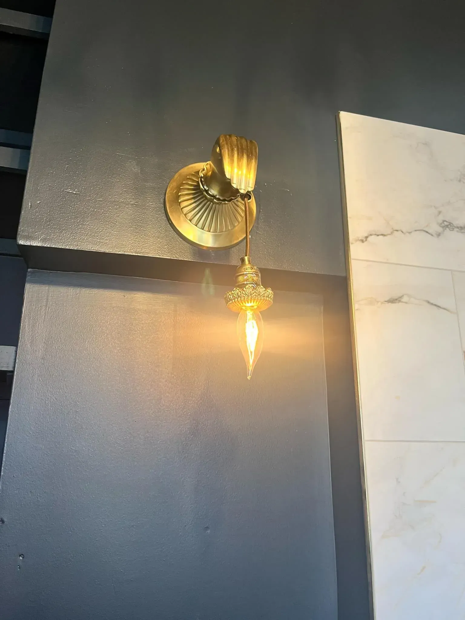 Gold sconce with a pointed bulb against a dark gray wall, next to a marble wall.