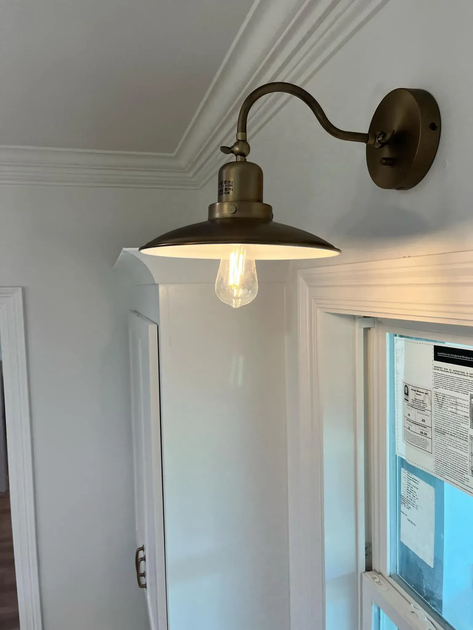 Brass wall-mounted light fixture above a window, illuminating a white trim and wall.