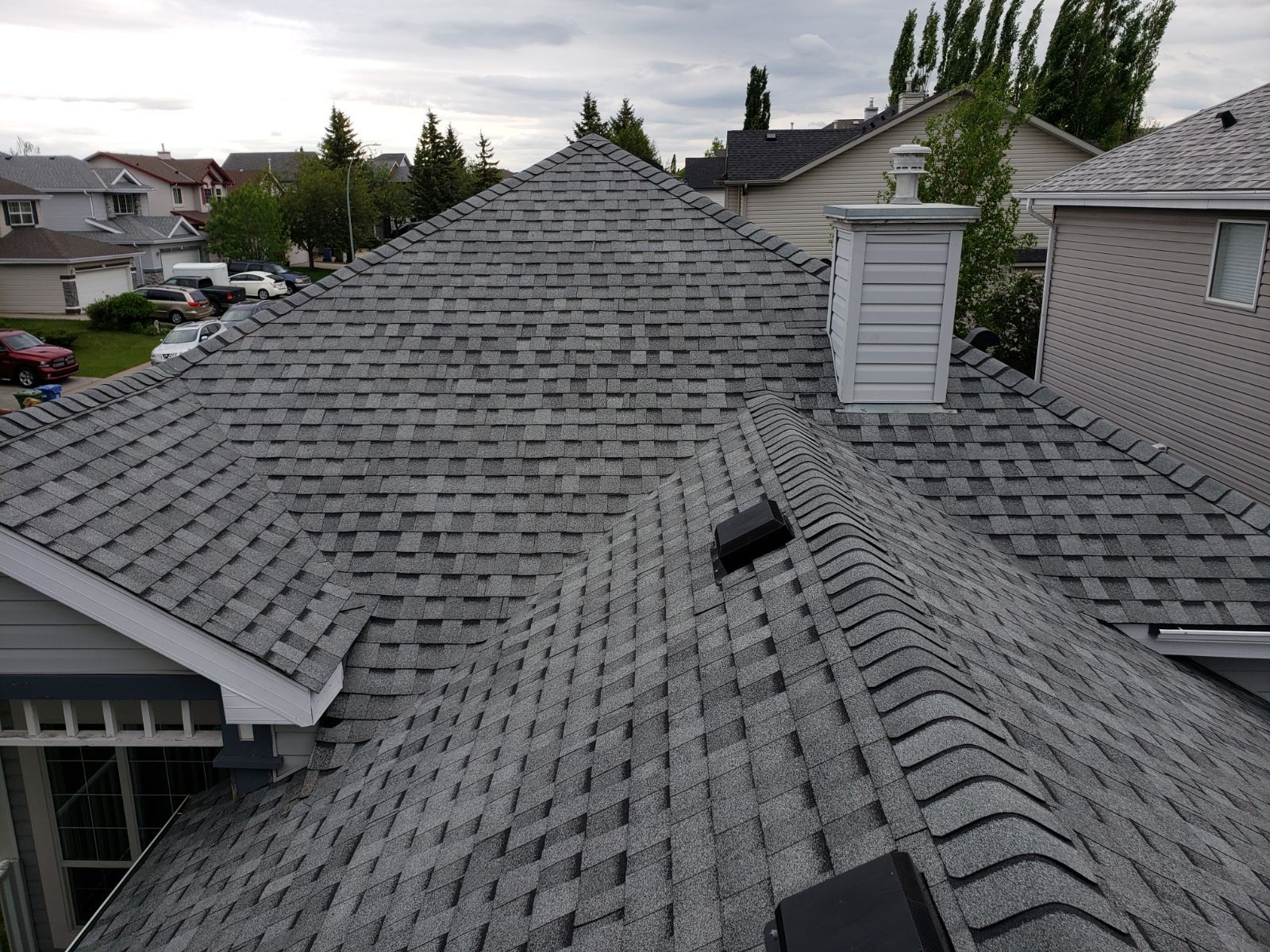 Gray asphalt shingle roof on a residential building with a chimney and small vent. Overcast sky.