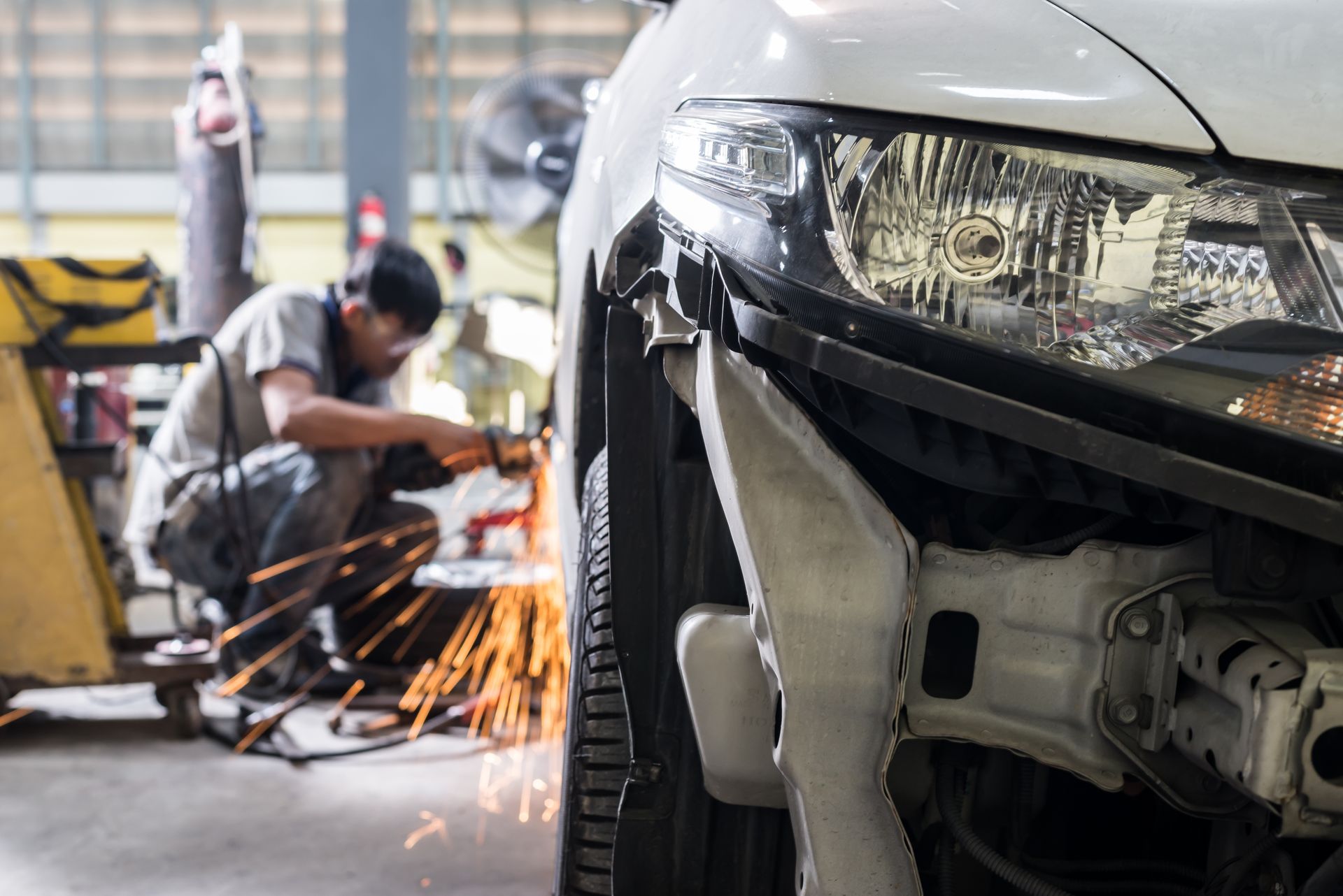 Car body repair scene with a worker welding near a damaged car bumper, sparks flying in a garage.