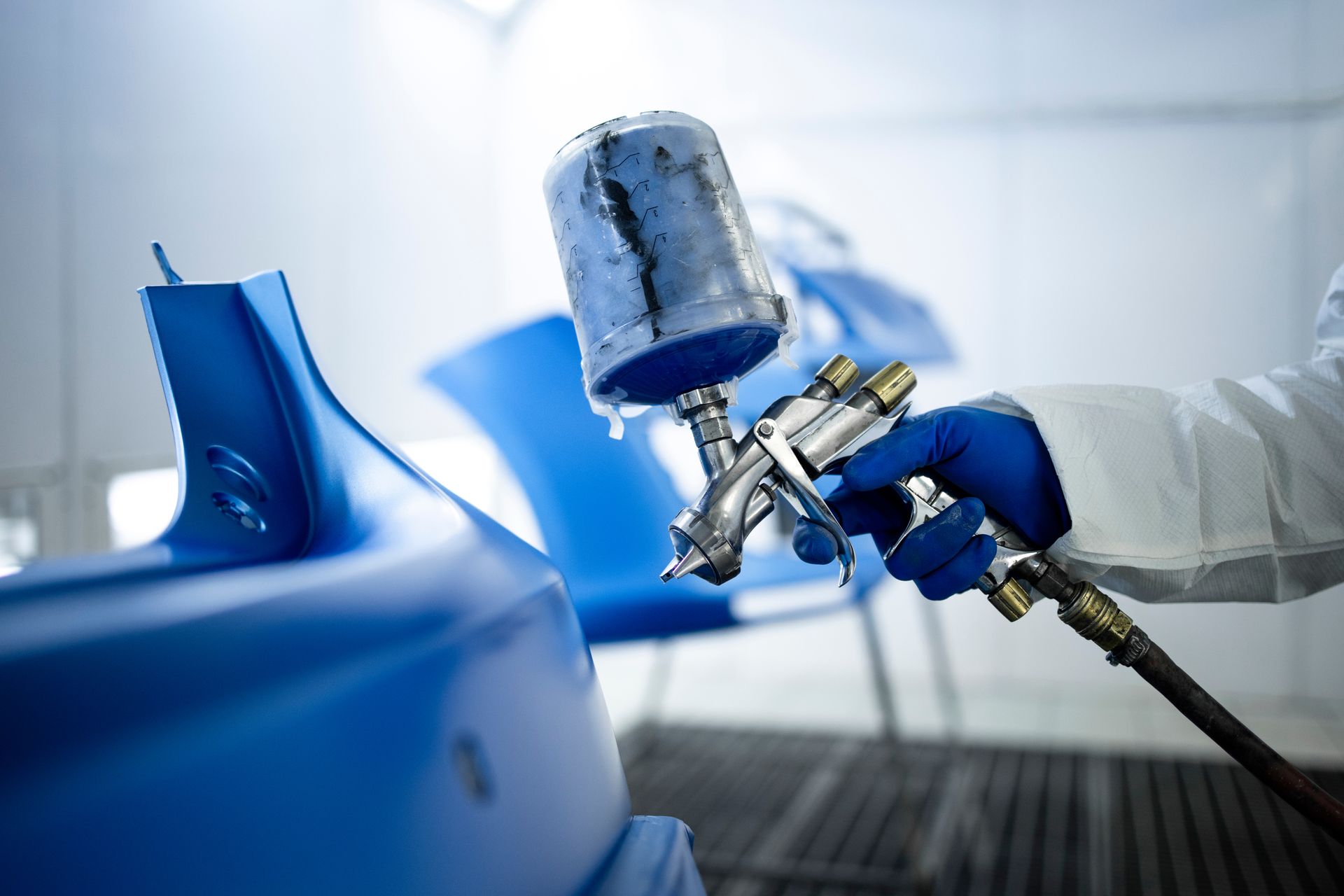 Blue car being spray-painted in a workshop by a gloved worker with a spray gun.