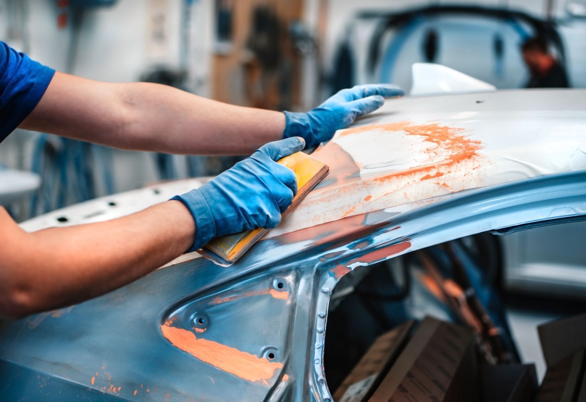 Gloved hands sanding a white car roof with orange body filler in an auto repair shop