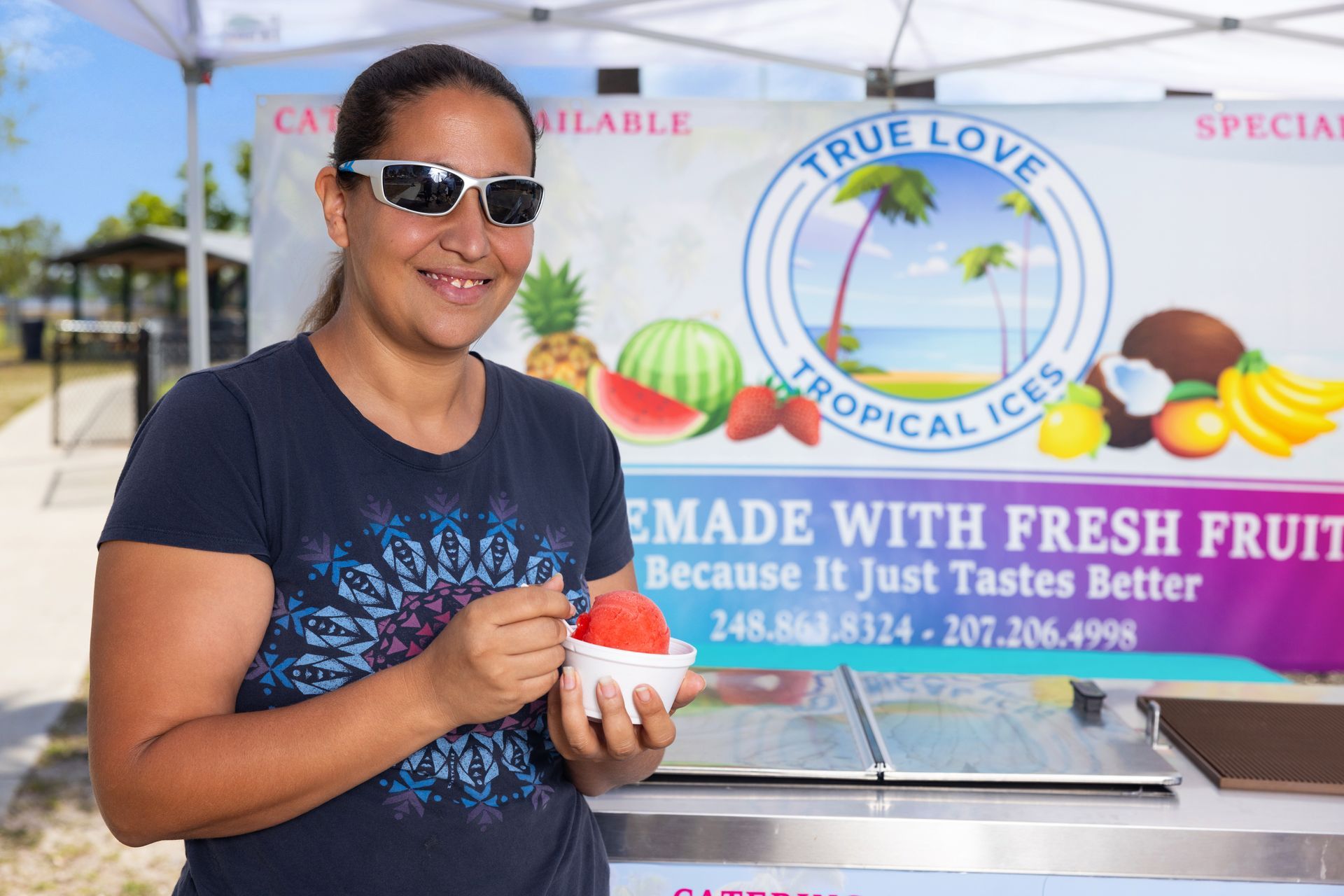 A woman is holding a cup of ice cream in front of a sign that says true love tropical ices