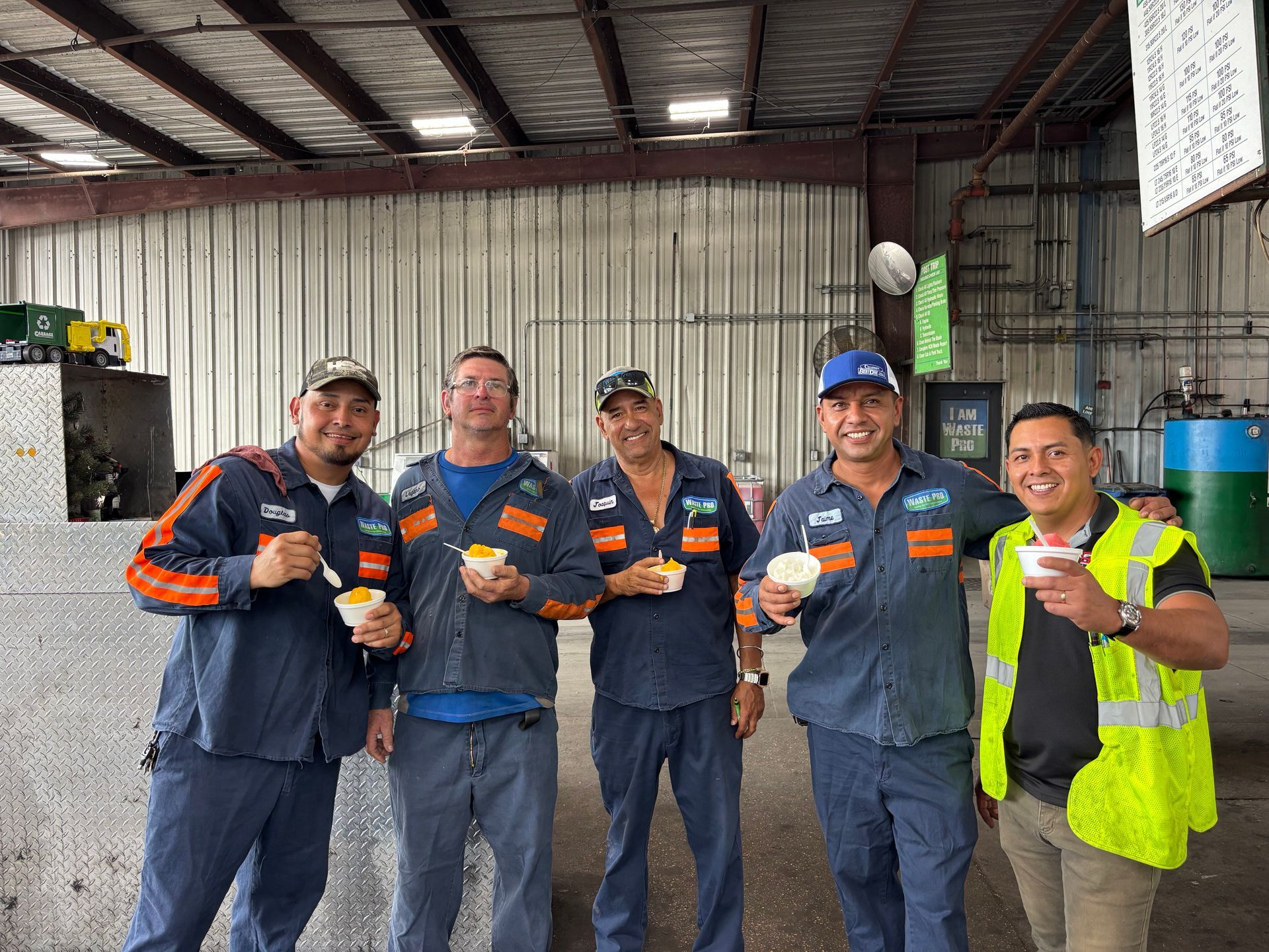 Five men in work uniforms smiling, holding food, inside a warehouse setting.