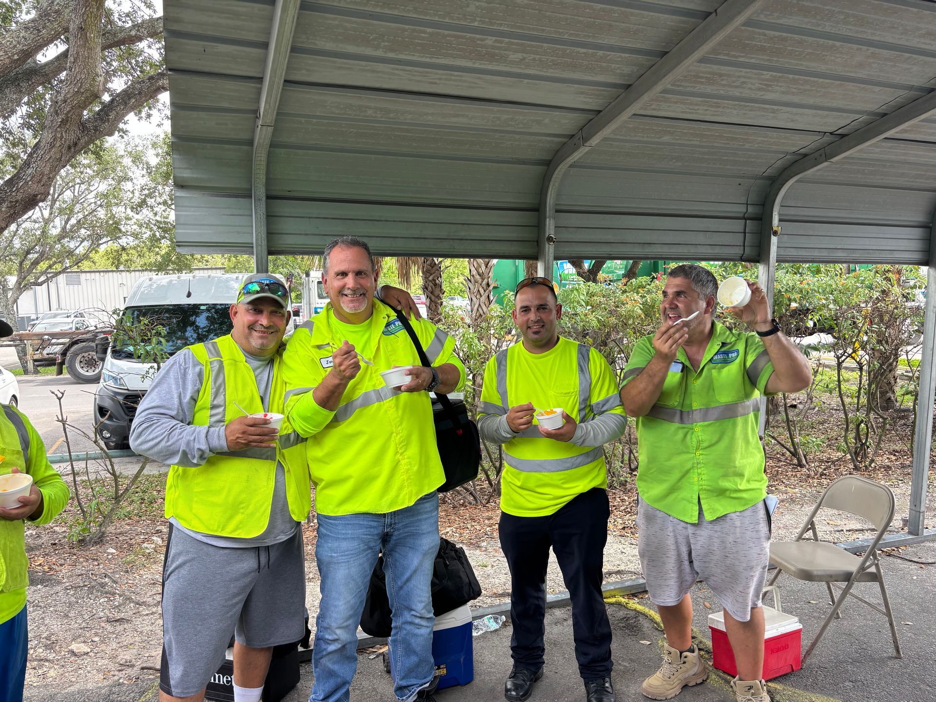 Four men in neon vests eating under a canopy; one man has food on his face.