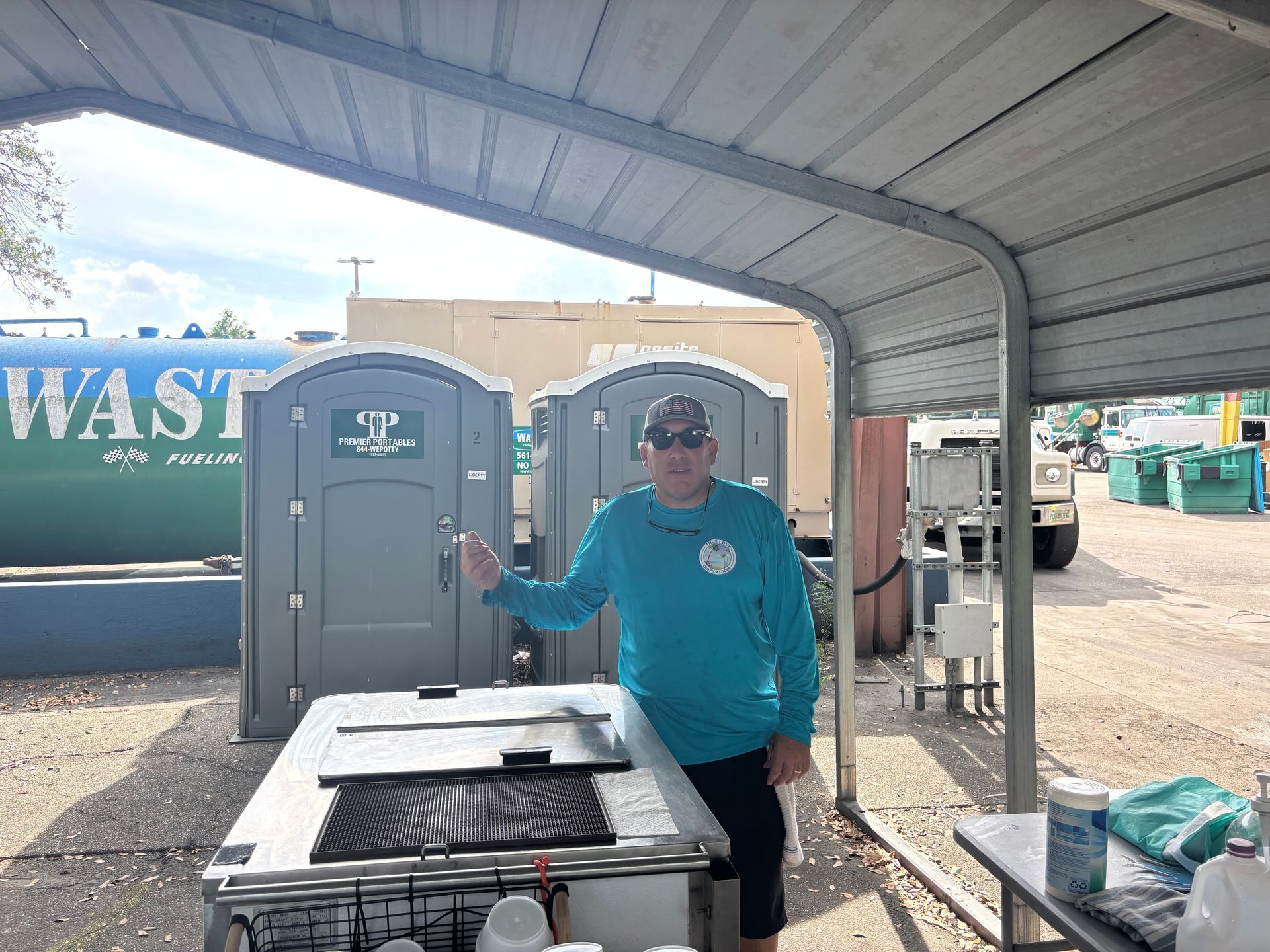 Man in blue shirt stands by a grill under a shelter, near portable toilets and a water tank.
