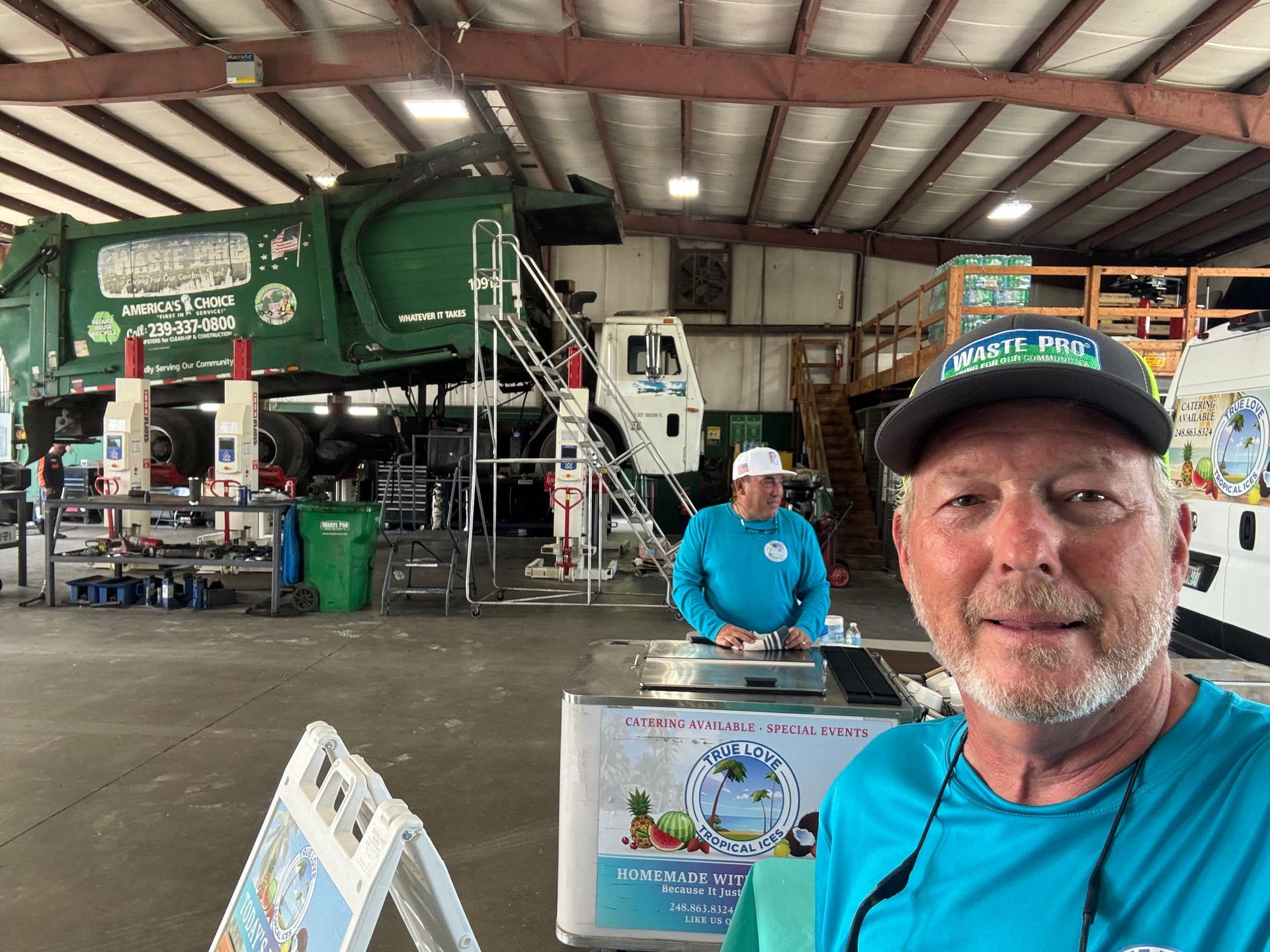 Man in teal shirt smiles, with an ice cream cart and a garbage truck being worked on in a garage.