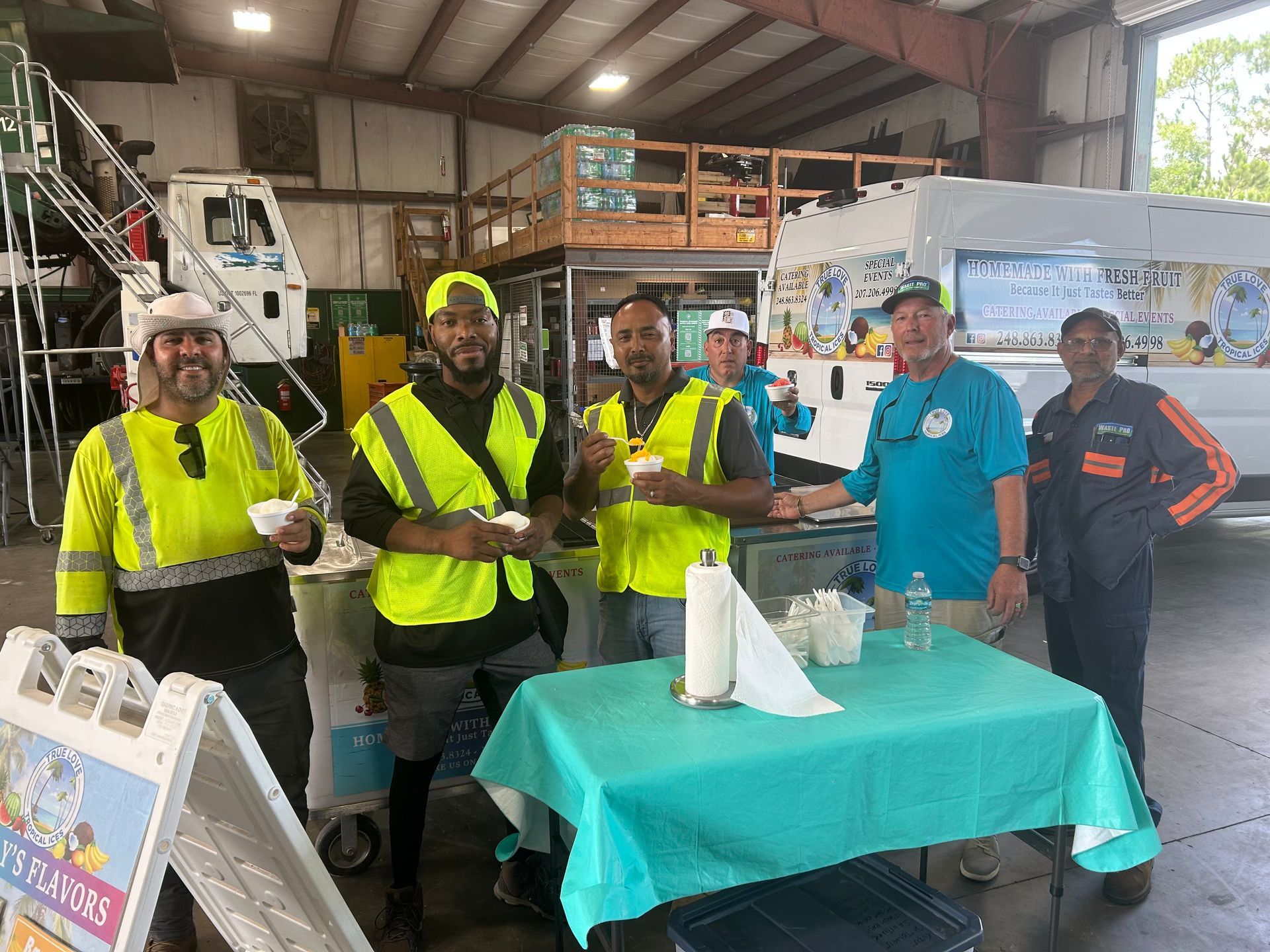 Group of workers at an outdoor gathering, some wearing high-vis vests, eating food near a table.