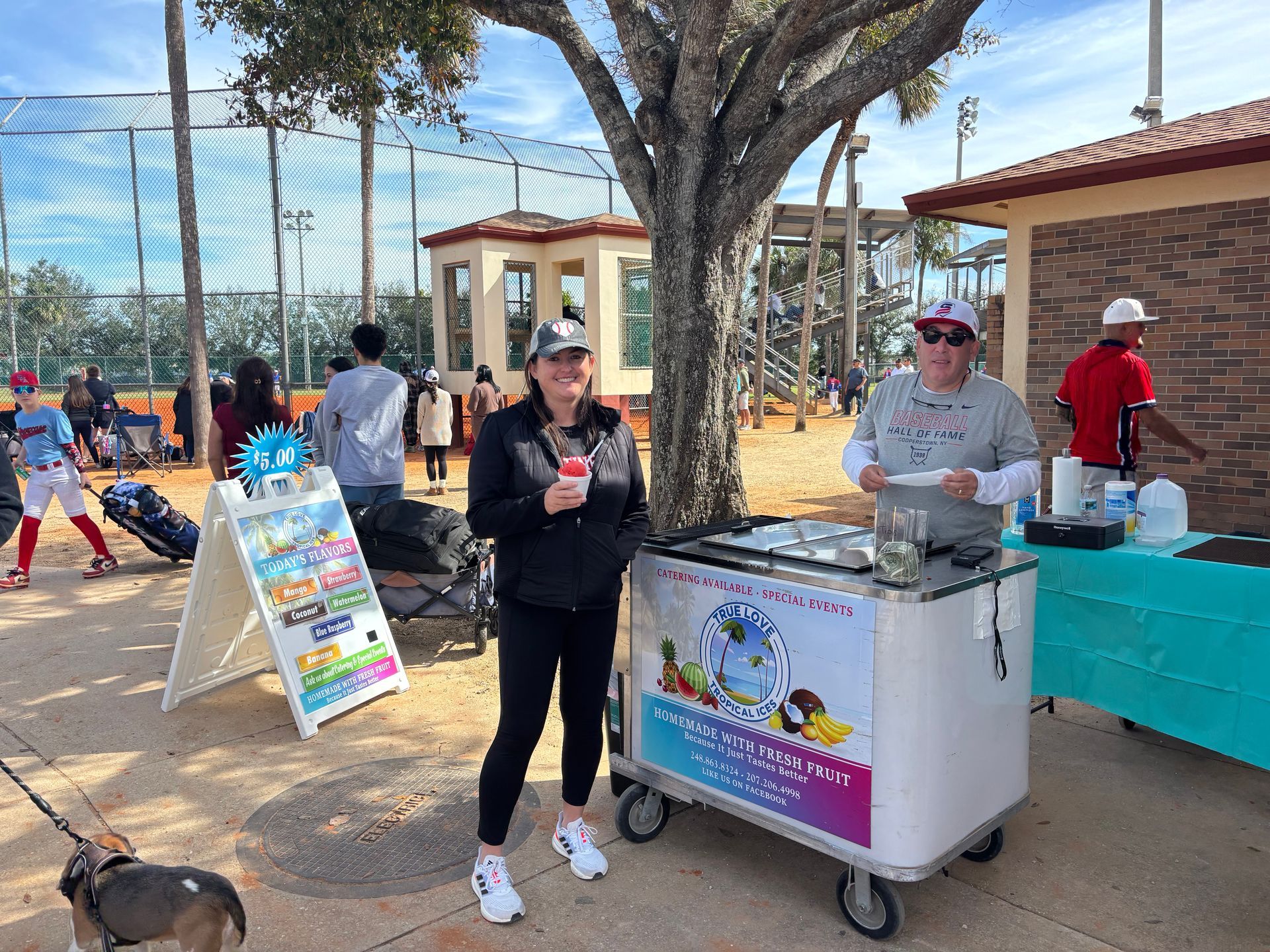 Two women are standing next to an ice cream cart in a park.