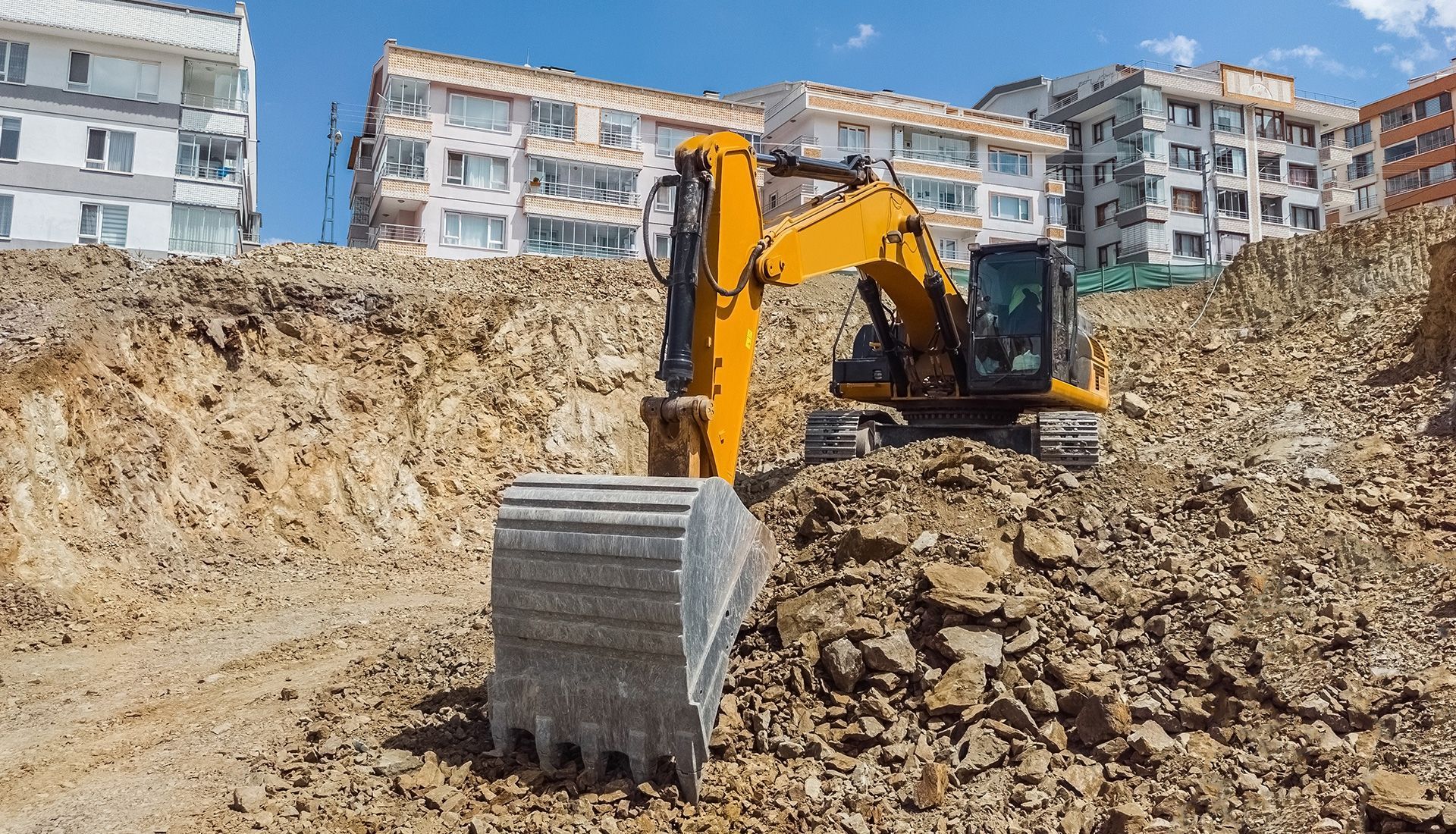 Yellow excavator digging into a hillside at a construction site; apartment buildings in the background.