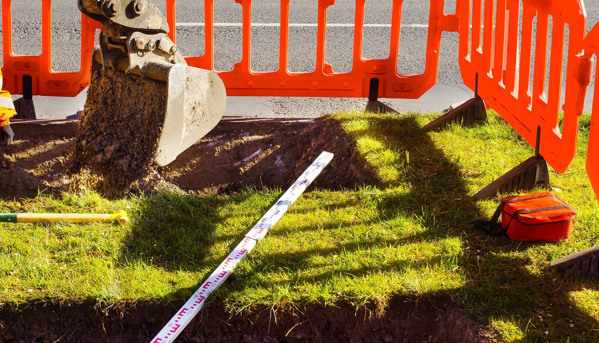 Excavator digging in grassy area, bordered by orange safety fencing.
