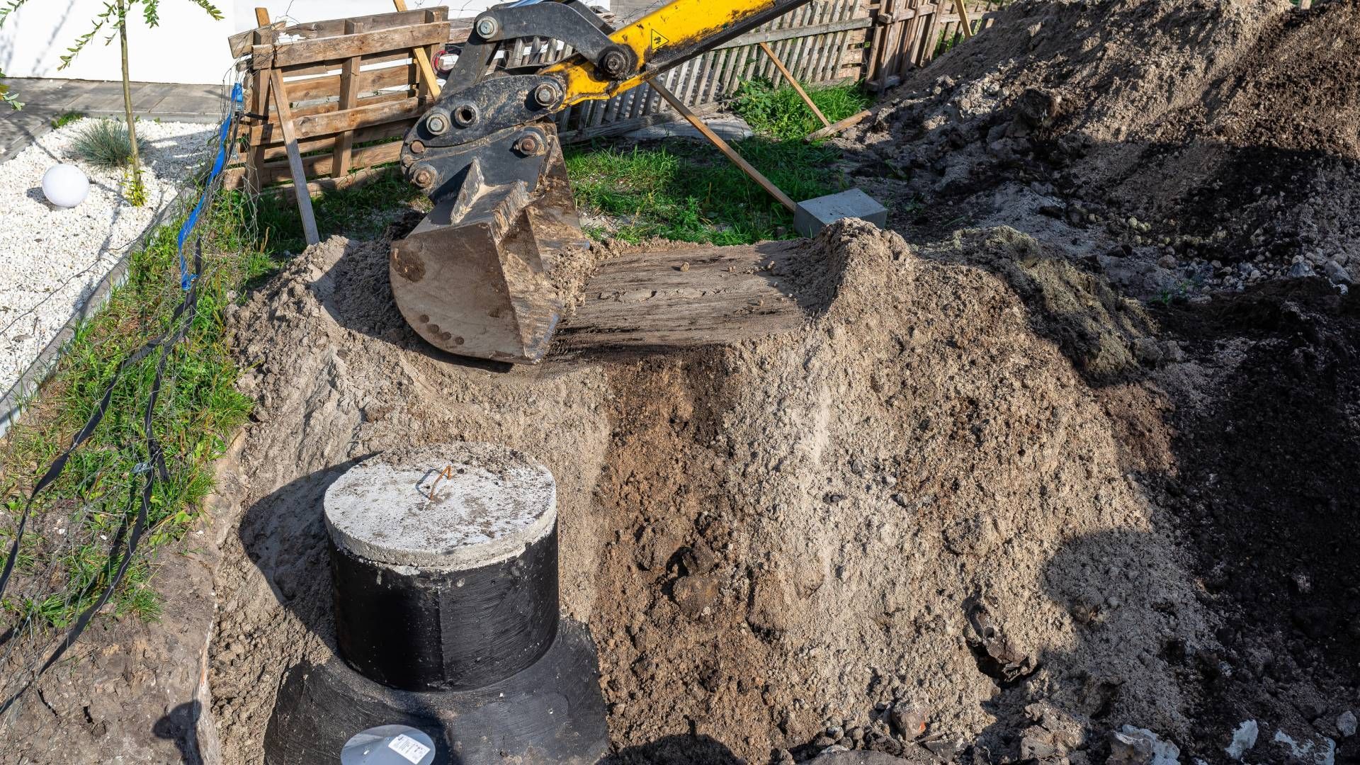 Excavator digging around a concrete septic tank in a yard.