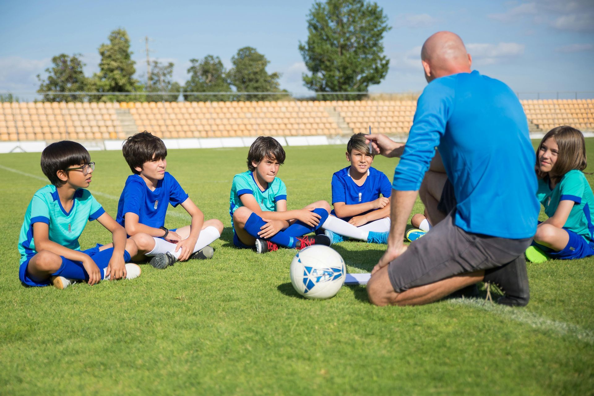 Soccer coach kneels, talking to a group of young players seated on a grassy field.