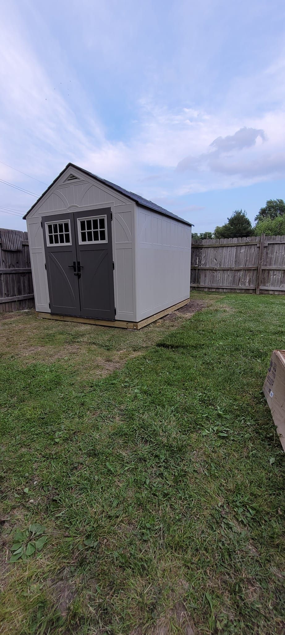 A small gray shed in a yard, with a dark roof and doors, under a cloudy sky.