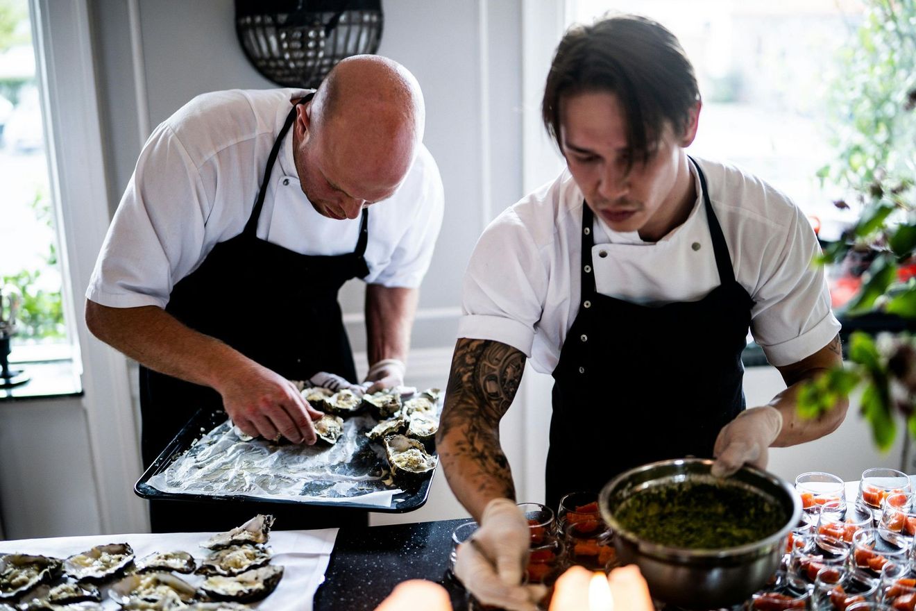Two chefs preparing food, one with tattoos, in a well-lit kitchen. One adds a topping to oysters.