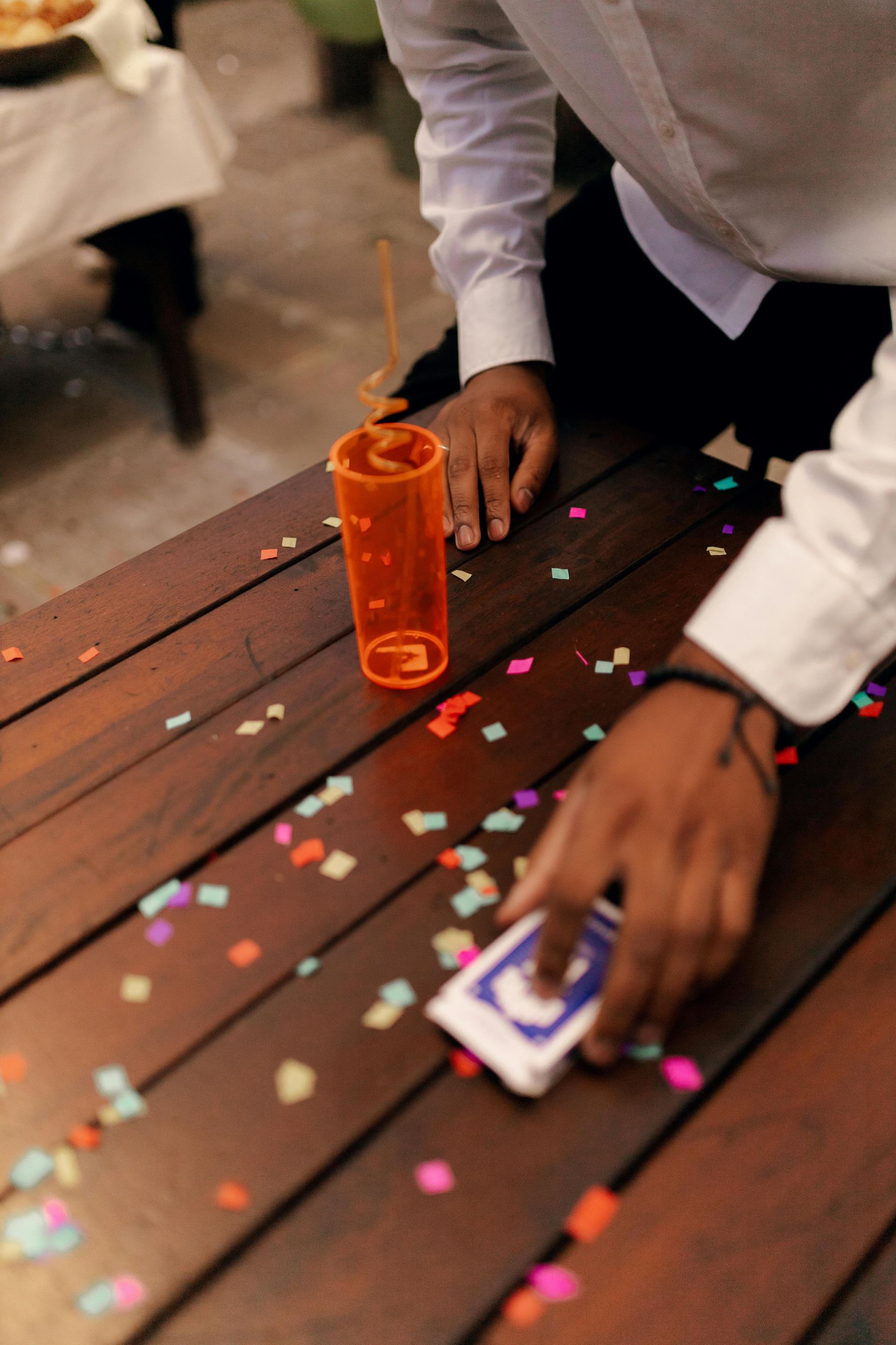 Person’s hands on a wooden table with confetti and a drink, cleaning up with a deck of cards.