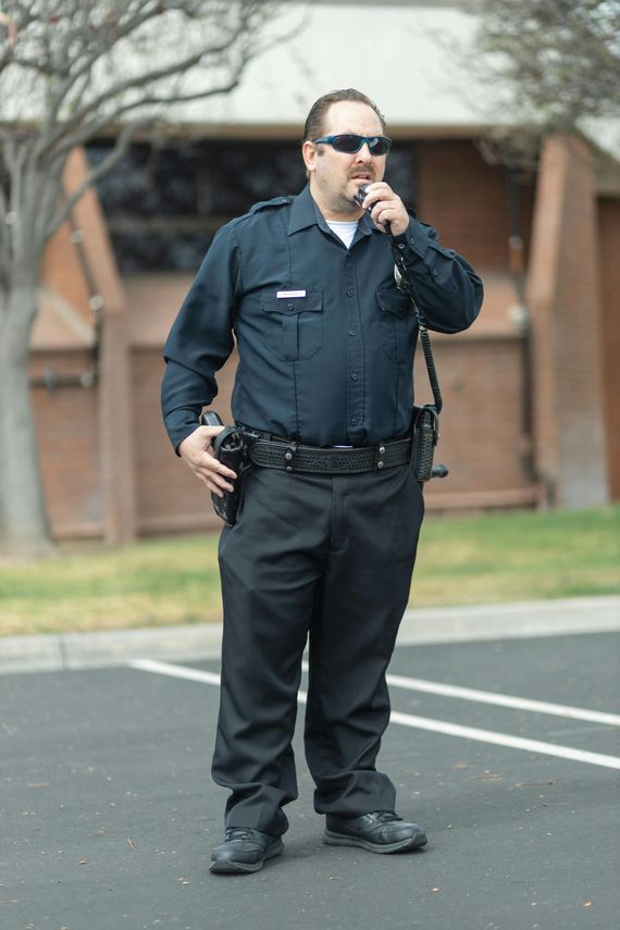 Man in a dark uniform and sunglasses holding a radio in a parking lot.
