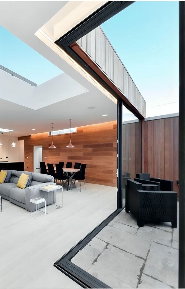 Modern living room with open sliding glass door to patio. Features wood accents, a grey sofa, and dining area.