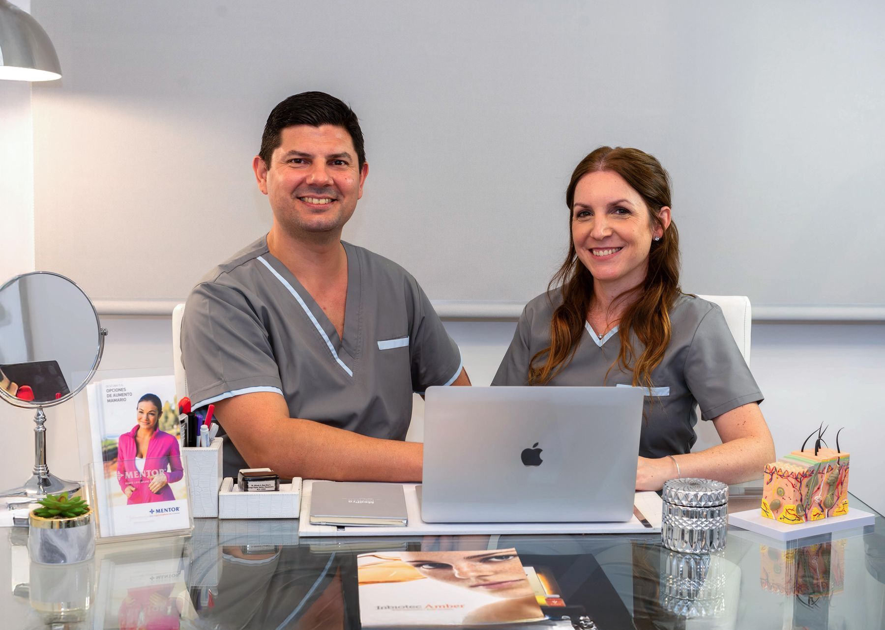 Dos personas con uniformes grises sonriendo ante un escritorio con una computadora portátil y una exhibición de productos para el cuidado de la piel.
