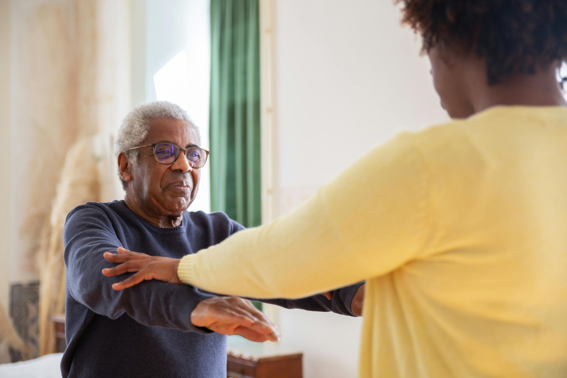 An older person stretches arms out with guidance from a person in a yellow shirt.
