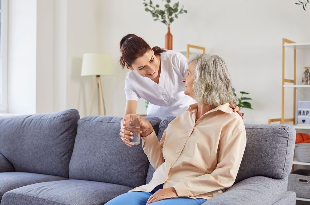Caregiver helping senior woman with a glass of water on a living room couch.