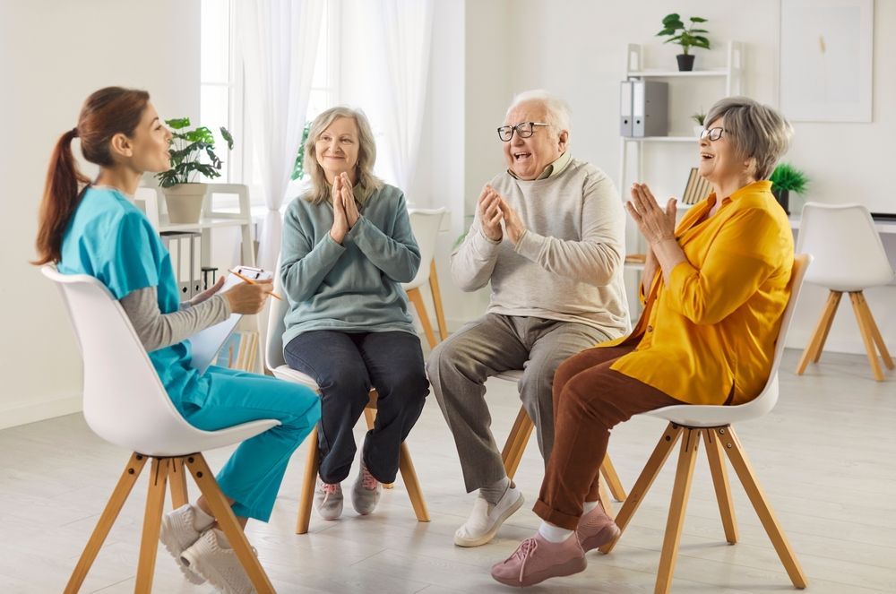 A caregiver and three seniors clapping in a bright room.