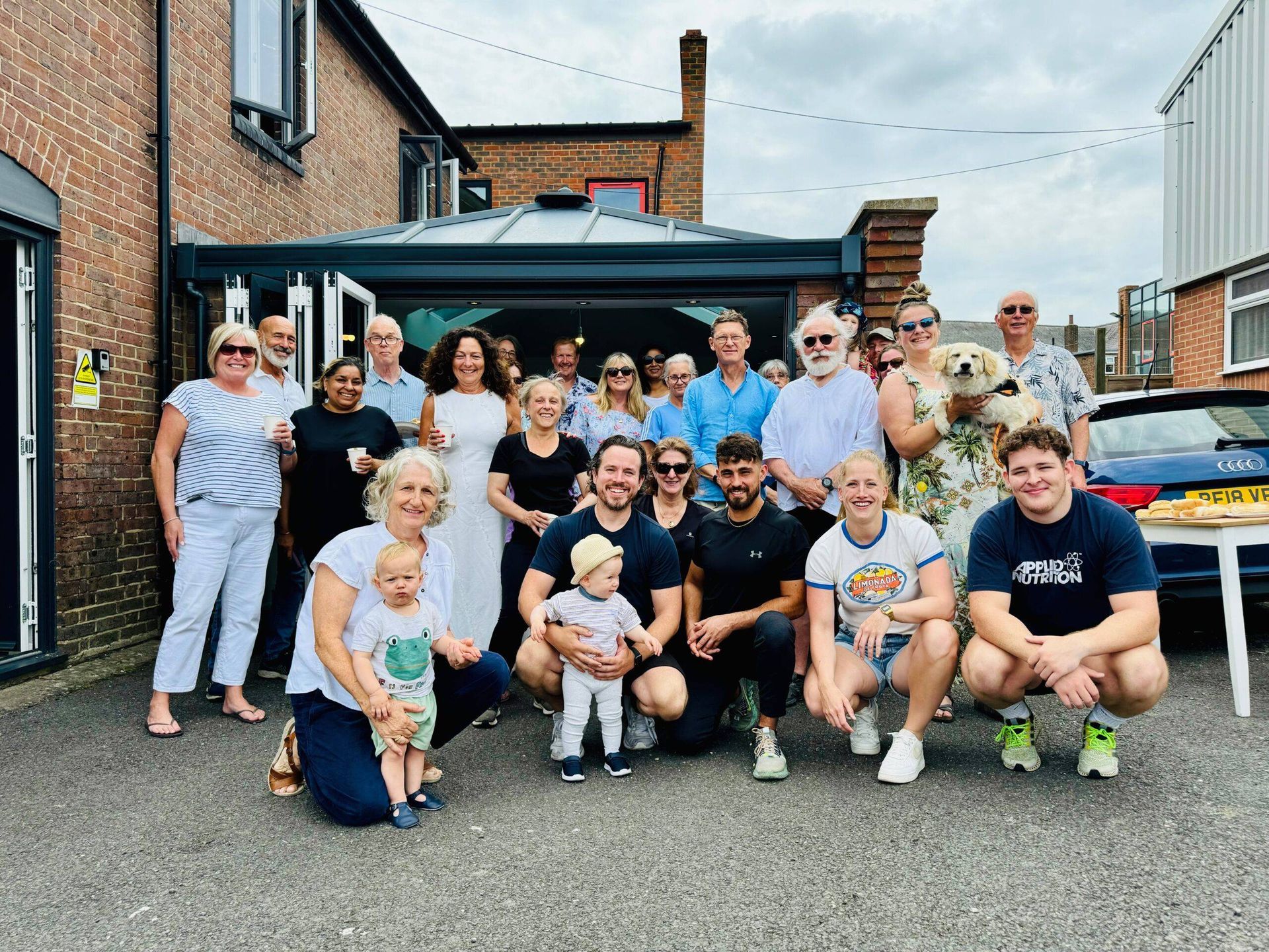 A group of people, of various ages, pose together in front of a brick building with a glass-walled back entrance.