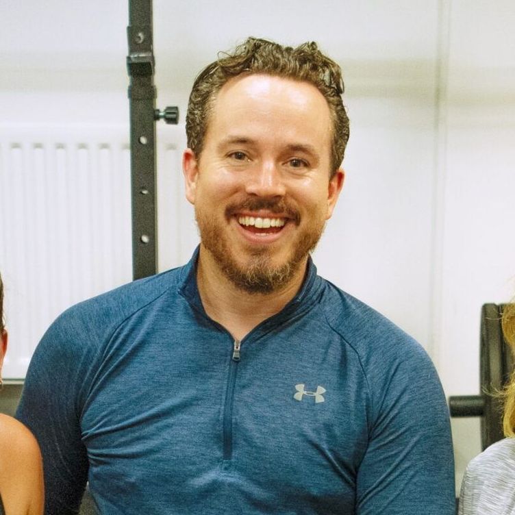 A smiling man with curly brown hair, wearing a blue Under Armour shirt, in a gym setting.