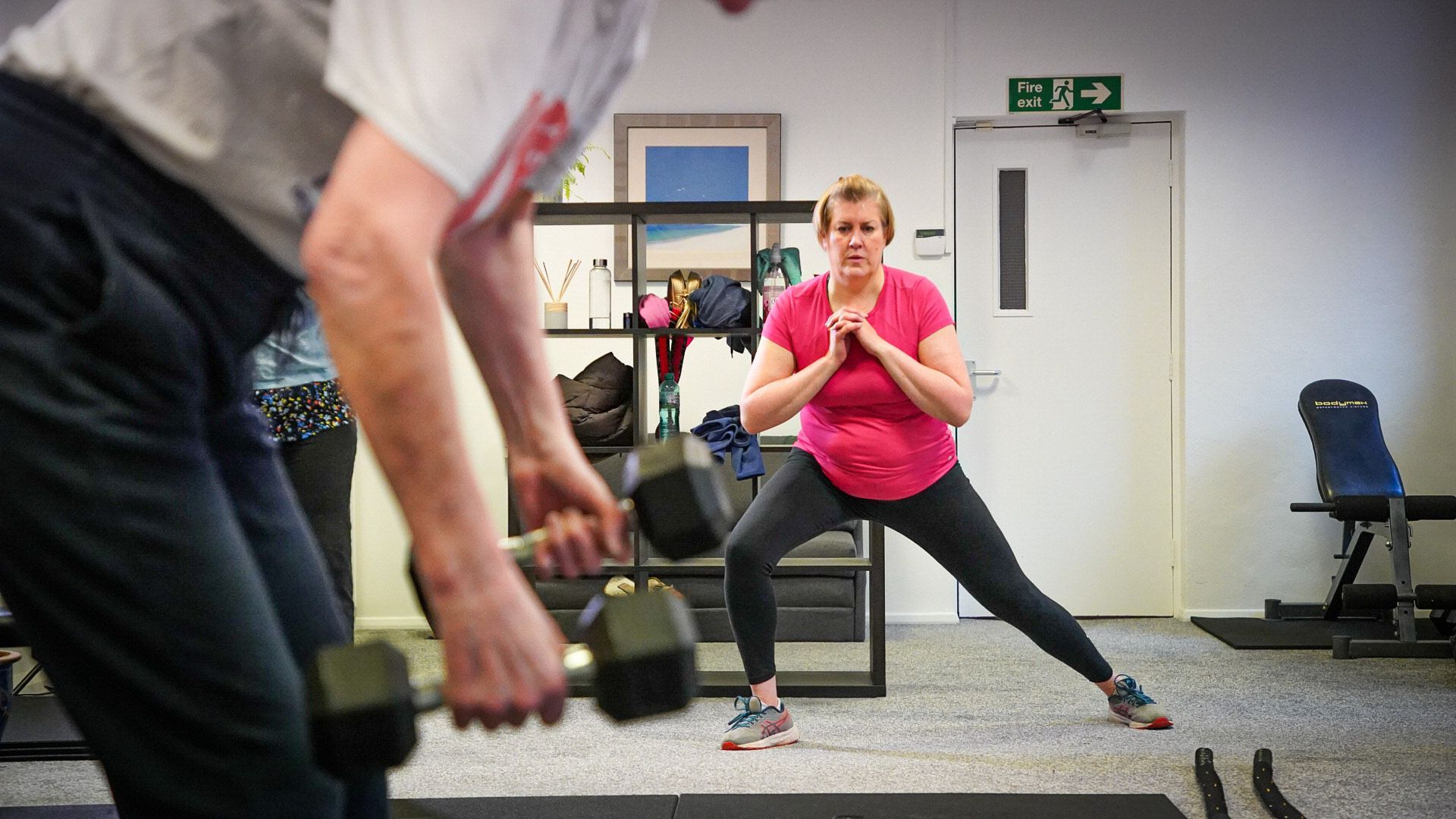 A woman does a side lunge exercise in a gym, while a man in the foreground holds dumbbells.