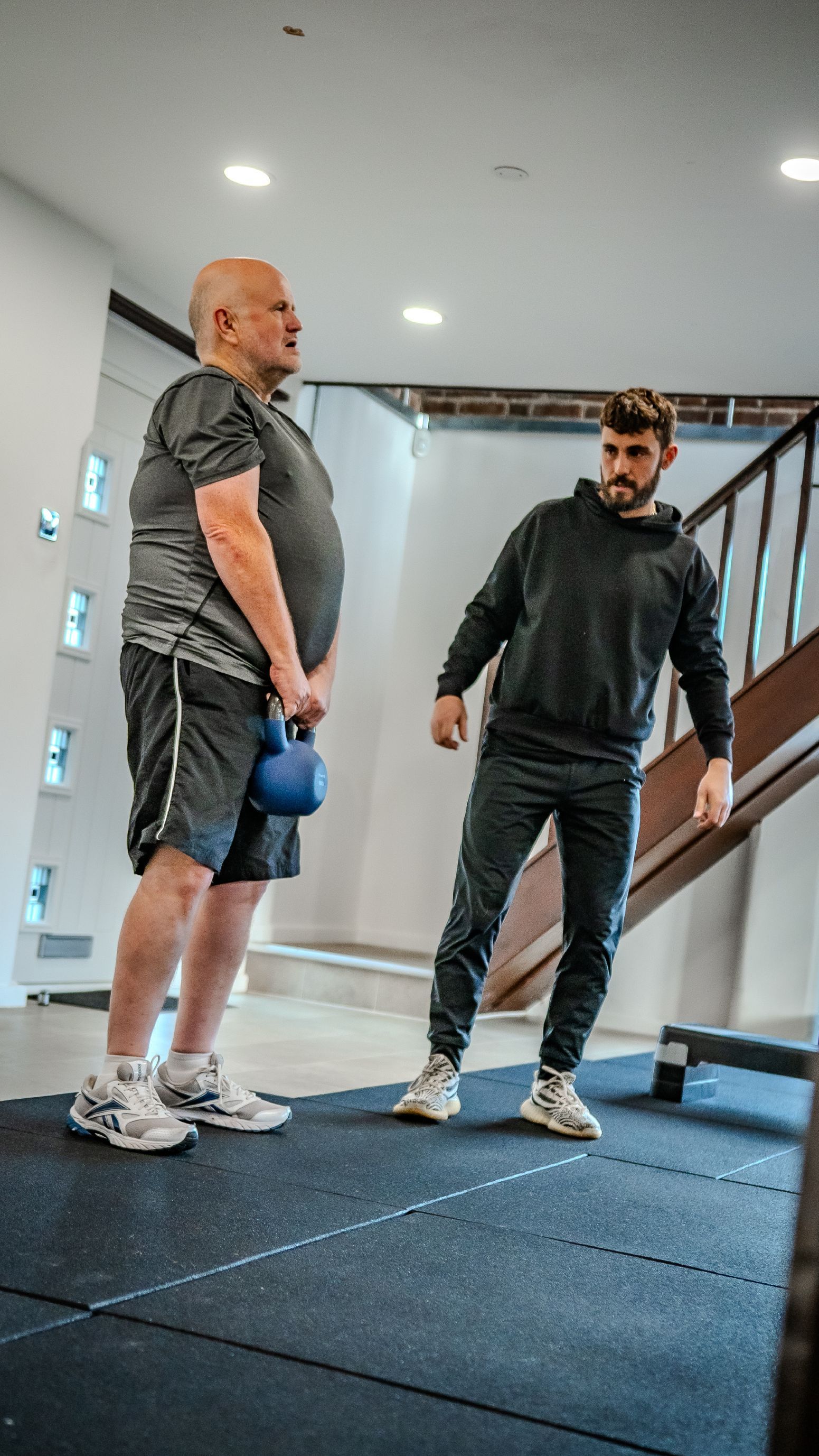 Two women in a gym setting: one doing step-ups while the other watches and smiles, offering guidance.