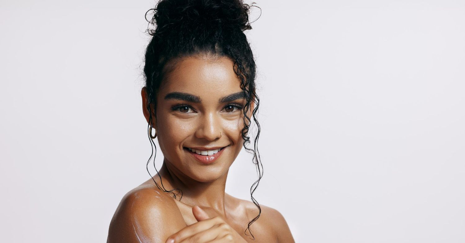 Woman with wet, curly hair in a bun smiles, touching her shoulder against a white backdrop.