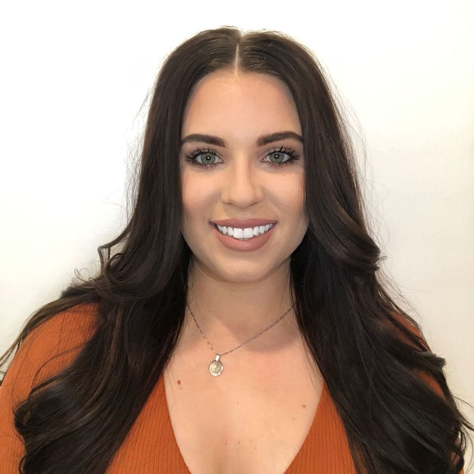 A woman wrapped in a white towel is smiling and touching her face.