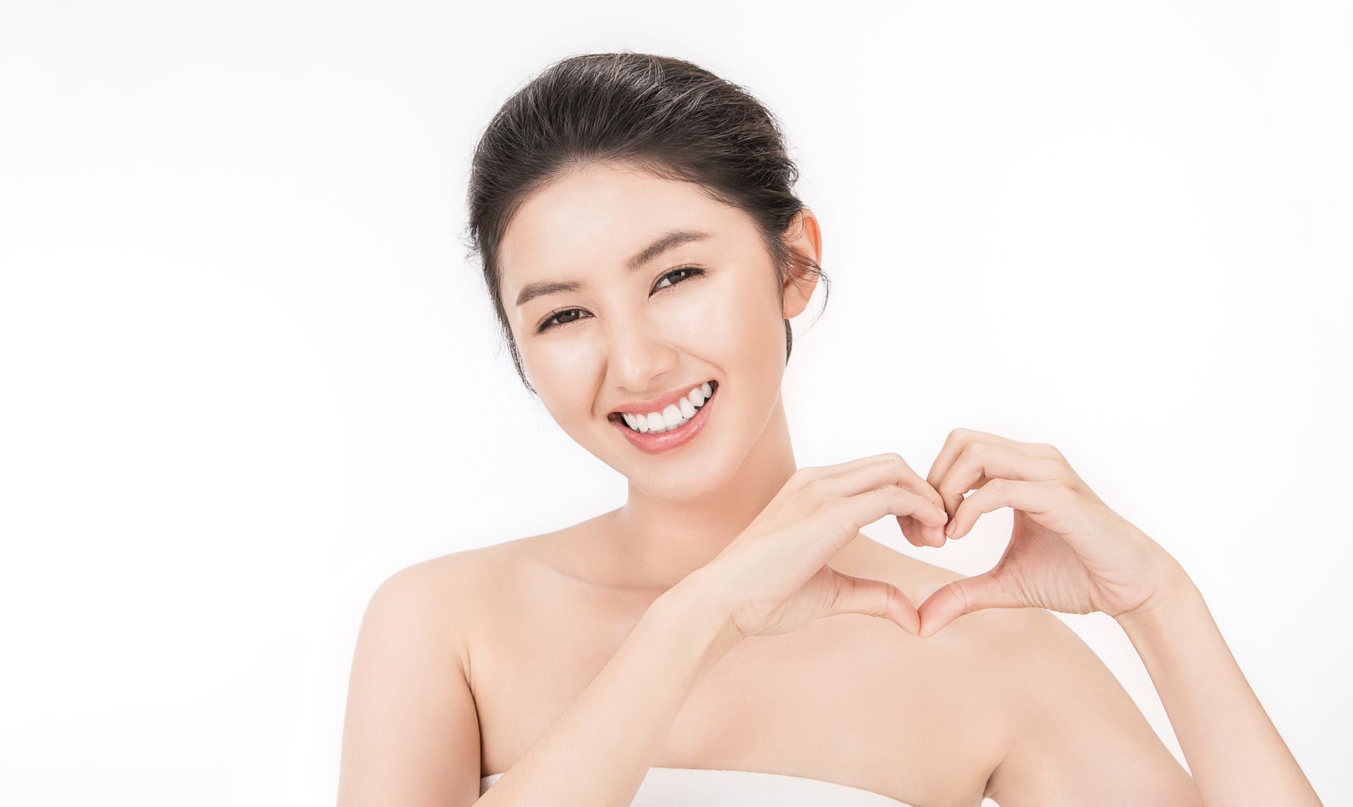 Woman smiles, forms heart with hands, white backdrop.